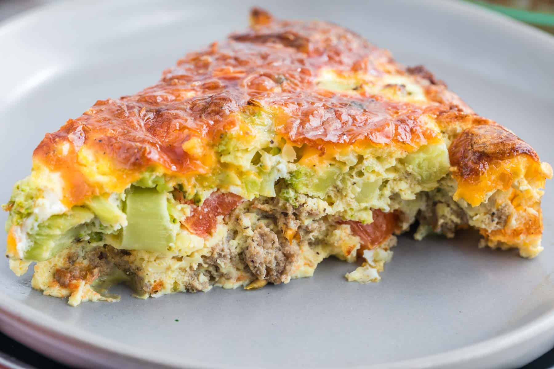 A close-up of a slice of quiche showing chunks of broccoli, tomato and sausage.