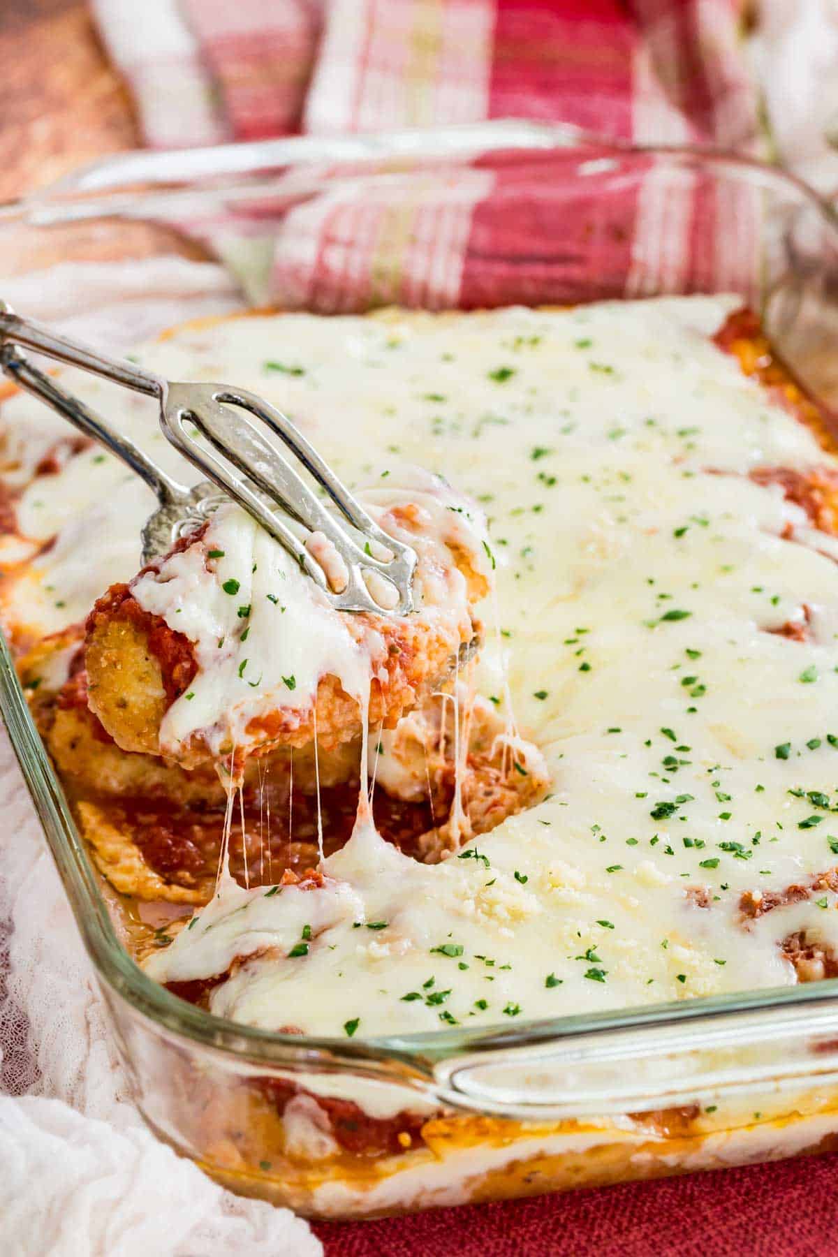 A piece of baked chicken is lifted from a glass casserole dish of cheesy chicken parmesan.