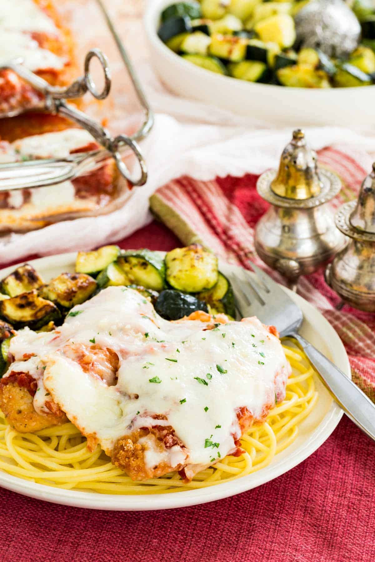 Gluten free chicken parmesan served over a plate of spaghetti with roasted zucchini on the side, with a casserole dish and bowl of zucchini in the background.