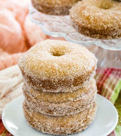 Three gluten free apple cider donuts stacked on a plate with more donuts on a cake stand in the background.