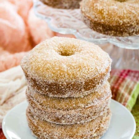 Three gluten free apple cider donuts stacked on a plate with more donuts on a cake stand in the background.