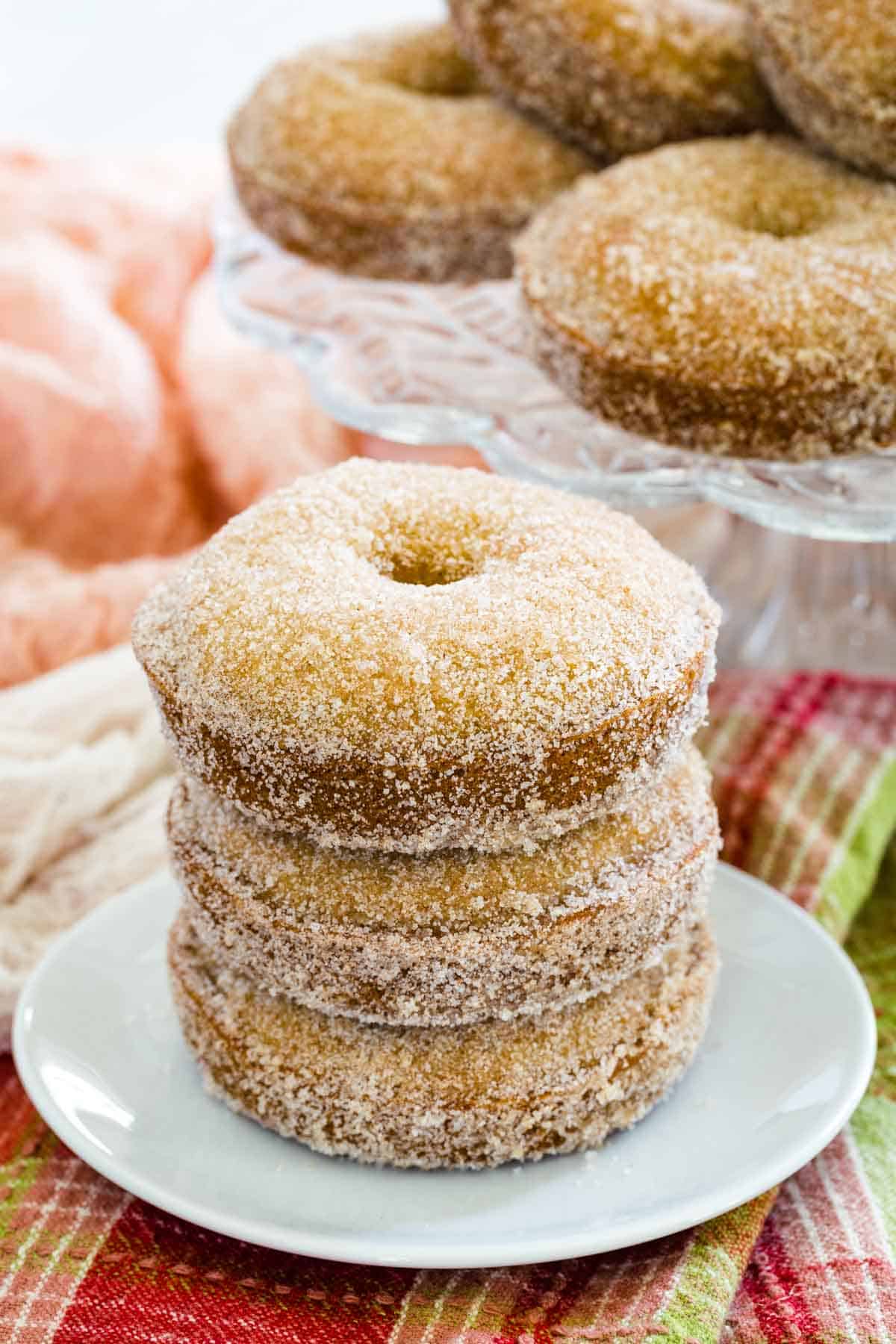 Three gluten free apple cider donuts stacked on a plate with more donuts on a cake stand in the background.