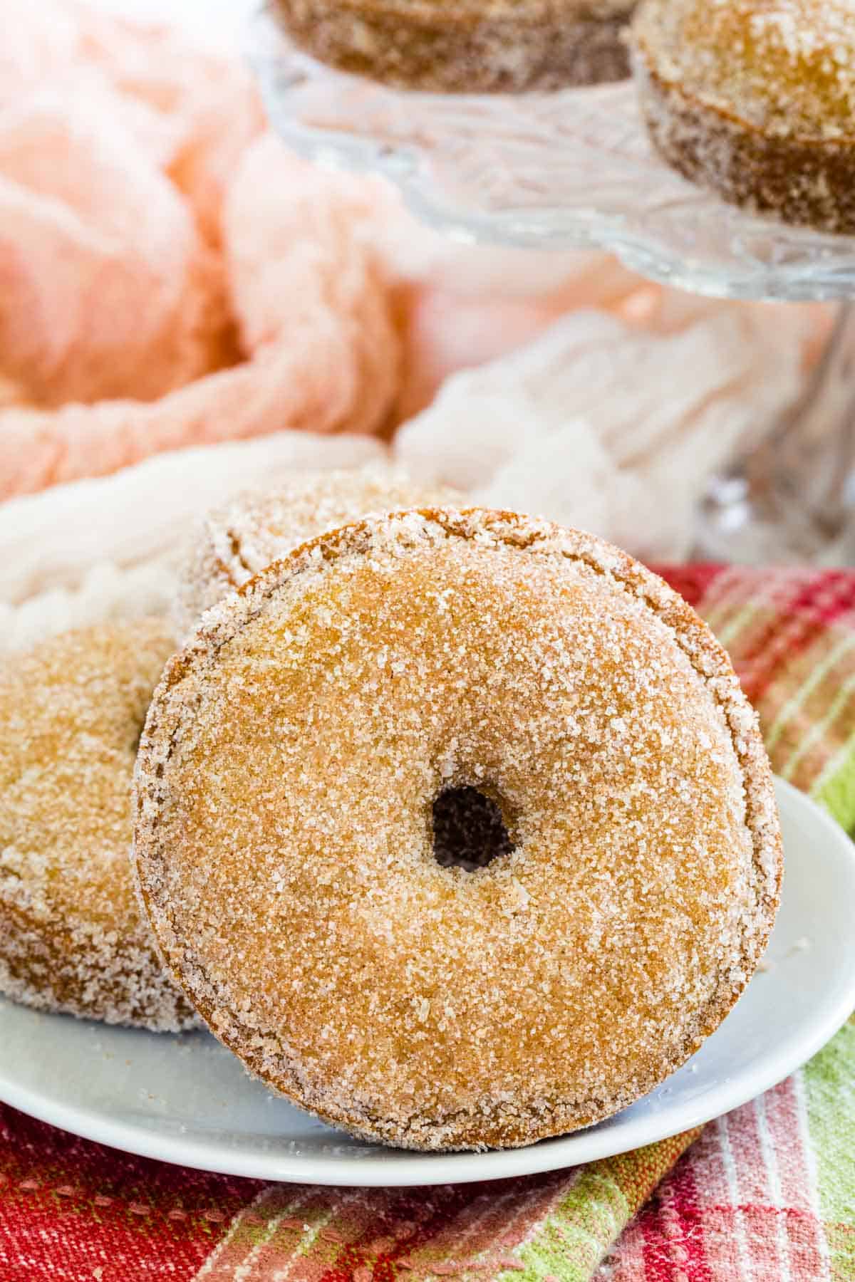 Close up of two gluten free apple cider donuts on a white plate, with more donuts on a glass cake stand in the background.