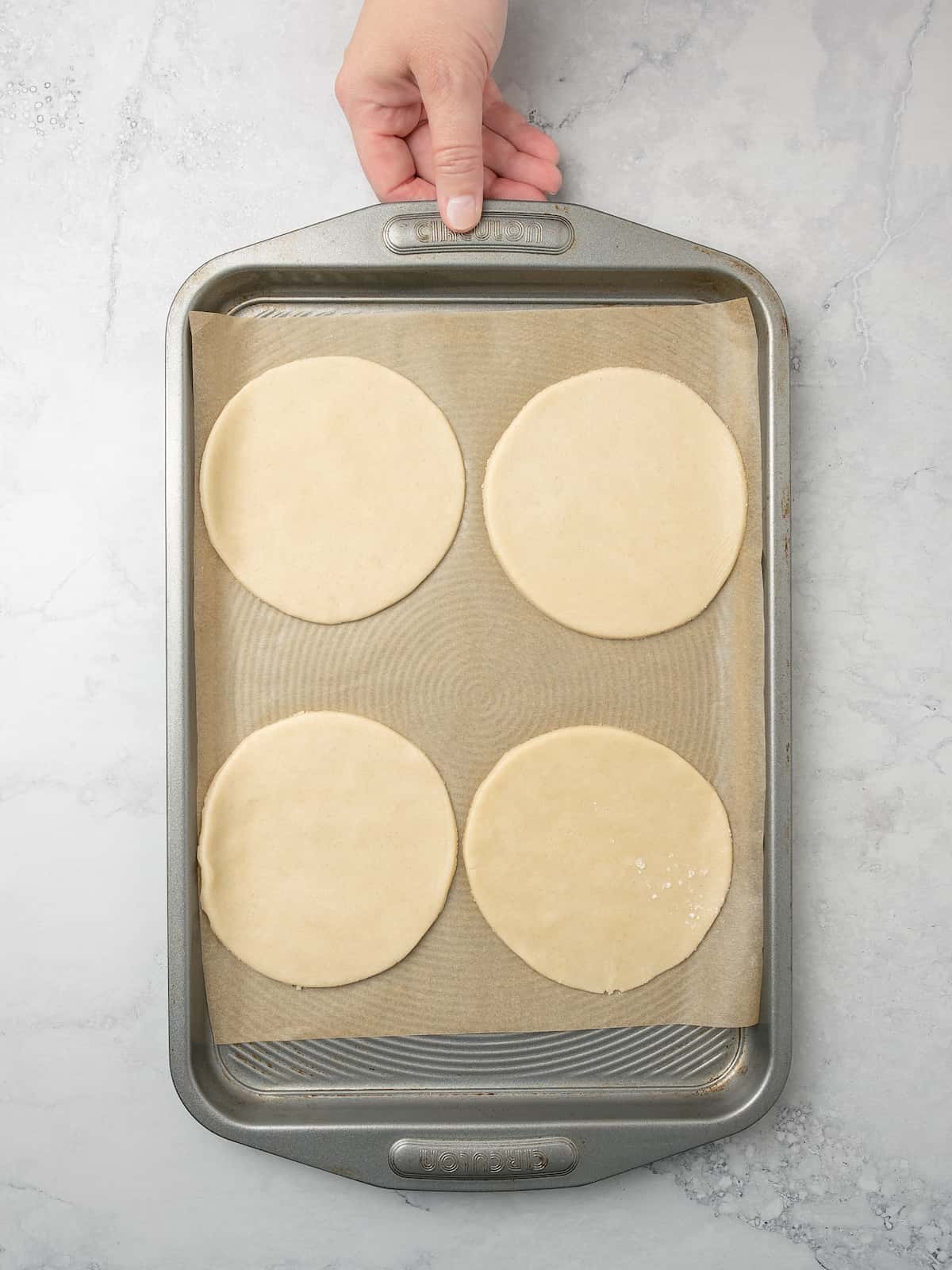 A hand holding a parchment lined baking sheet with four rounds of pastry dough.