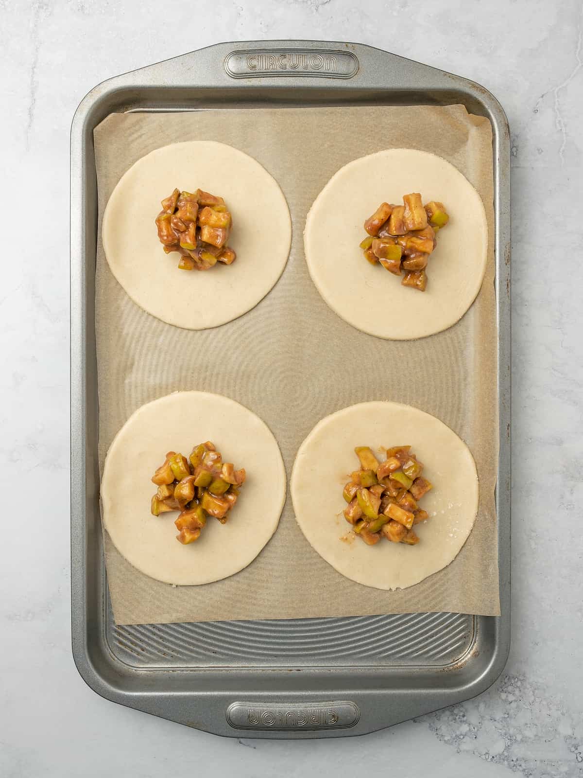 Four rounds of pastry dough topped with apple filling on a baking sheet lined with parchment paper.