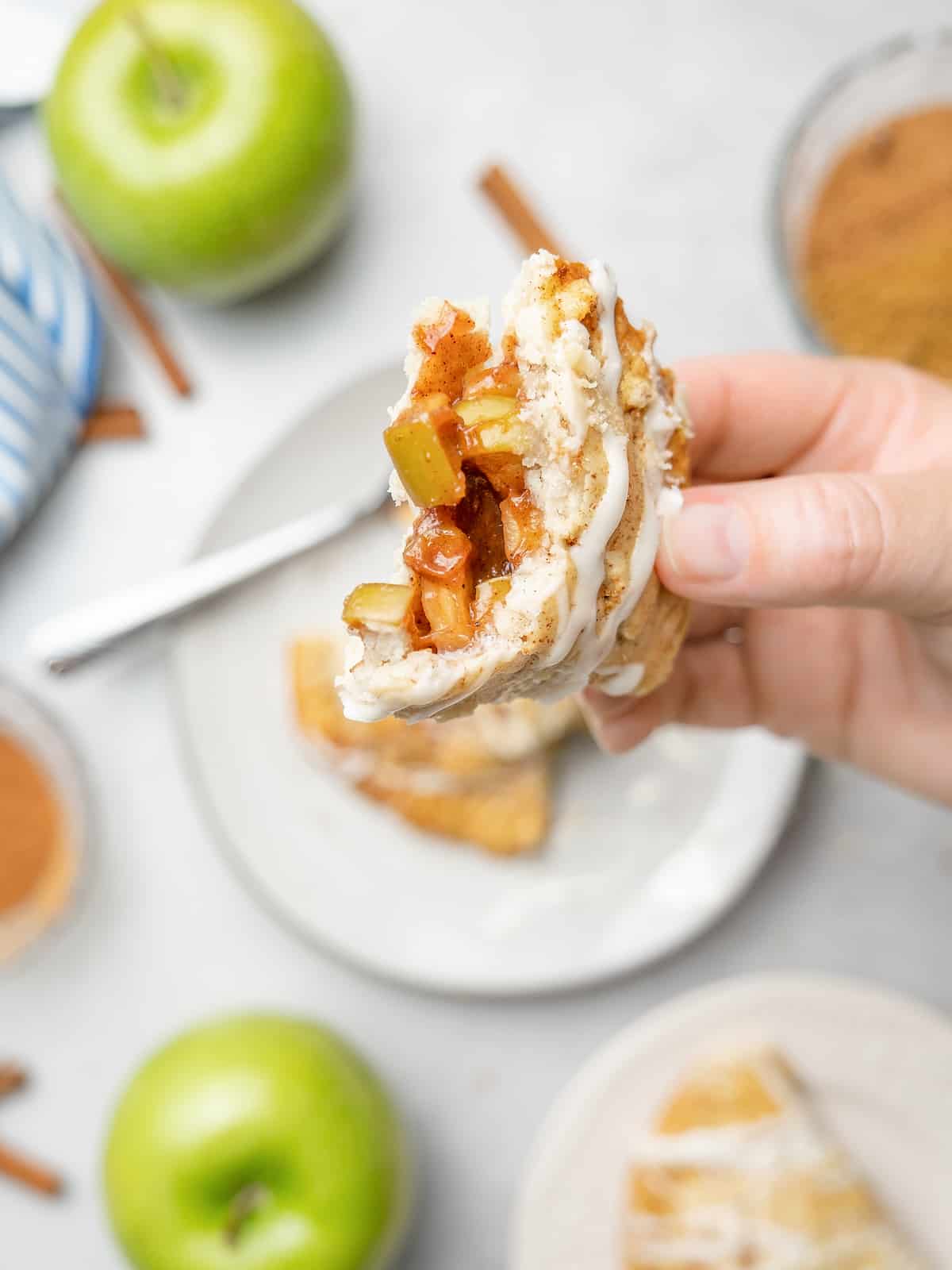 A hand holding an apple turnover that's been torn open to reveal the apple filling, with a plate and apples in the background.