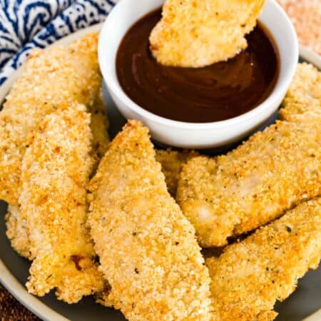 A hand dipping a crispy chicken tender into a small bowl of ketchup, on a plate surrounded by more baked chicken tenders.