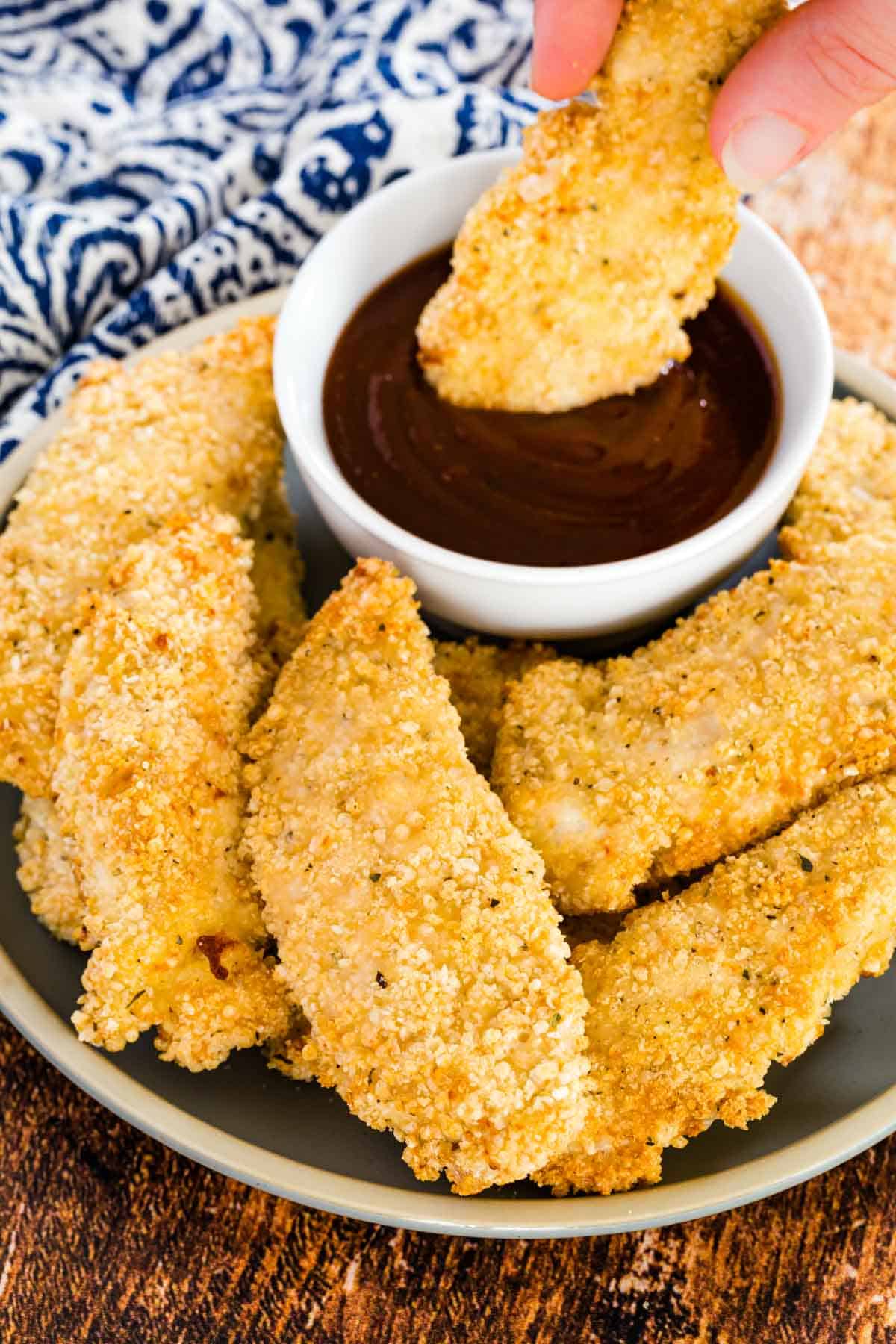 A hand dipping a crispy chicken tender into a small bowl of ketchup, on a plate surrounded by more baked chicken tenders.