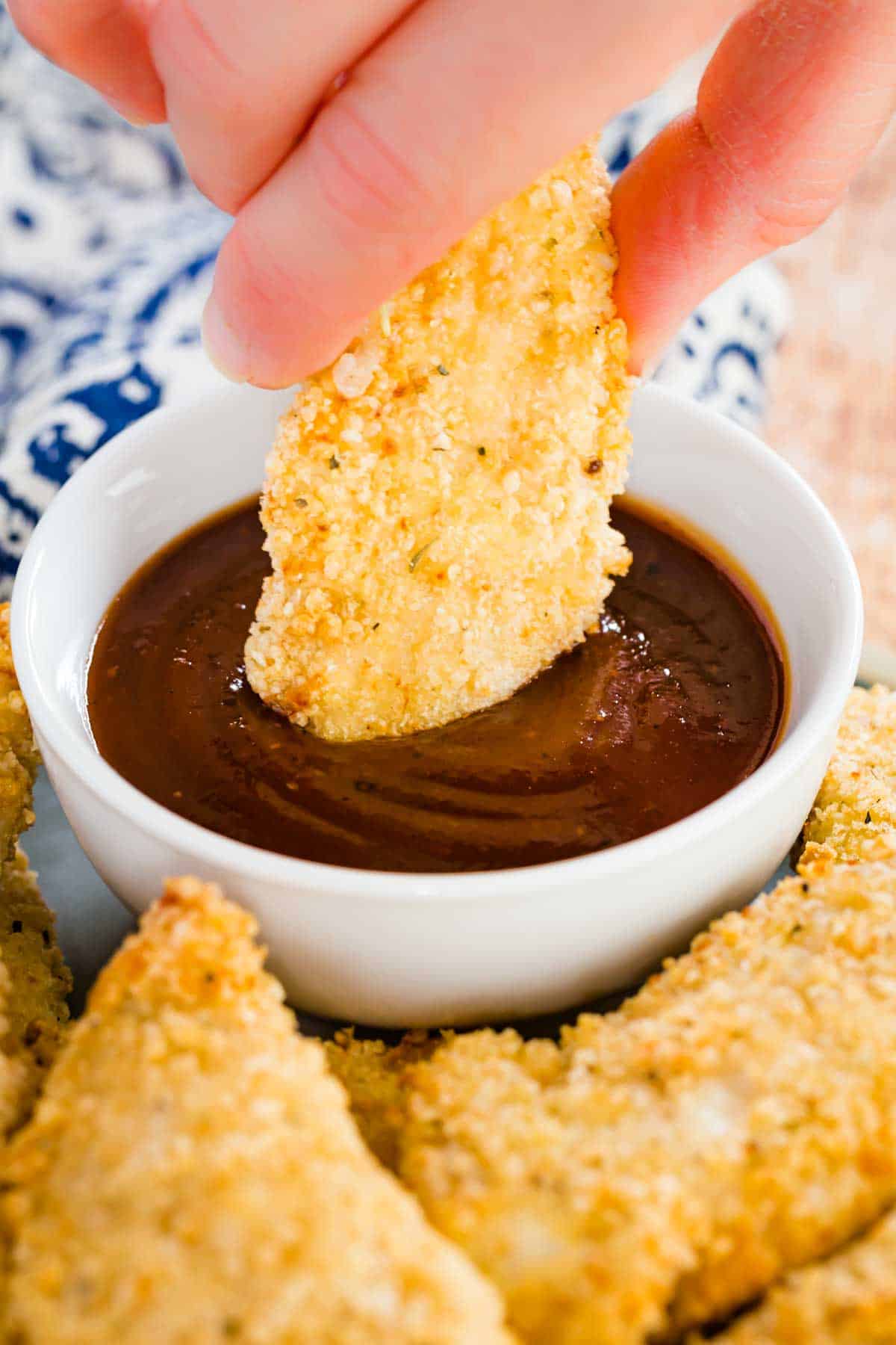 Close up of a hand dipping a crispy chicken tender into a small bowl of ketchup, on a plate surrounded by more baked chicken tenders.