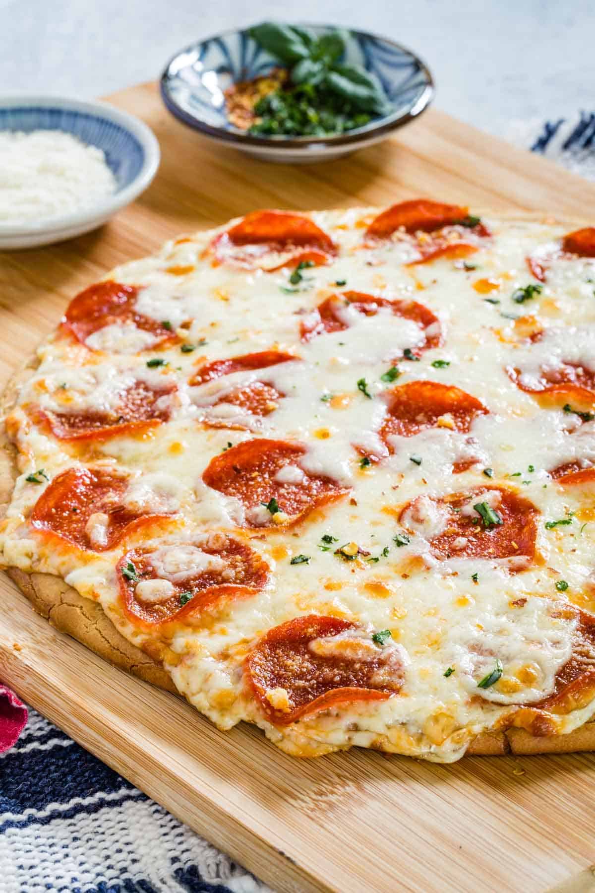 A whole gluten free pepperoni pizza on a wooden cutting board next to small bowls of basil and parmesan.