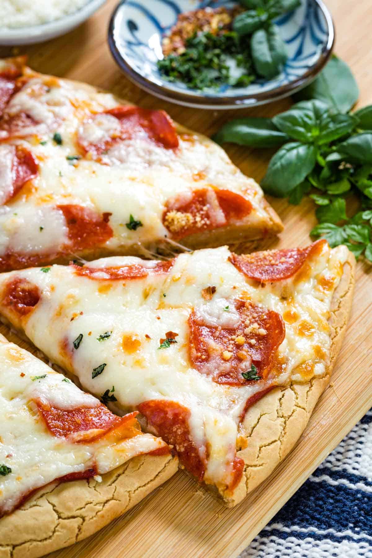 Gluten free pepperoni pizza slices on a wooden cutting board next to a small bowl of basil leaves.