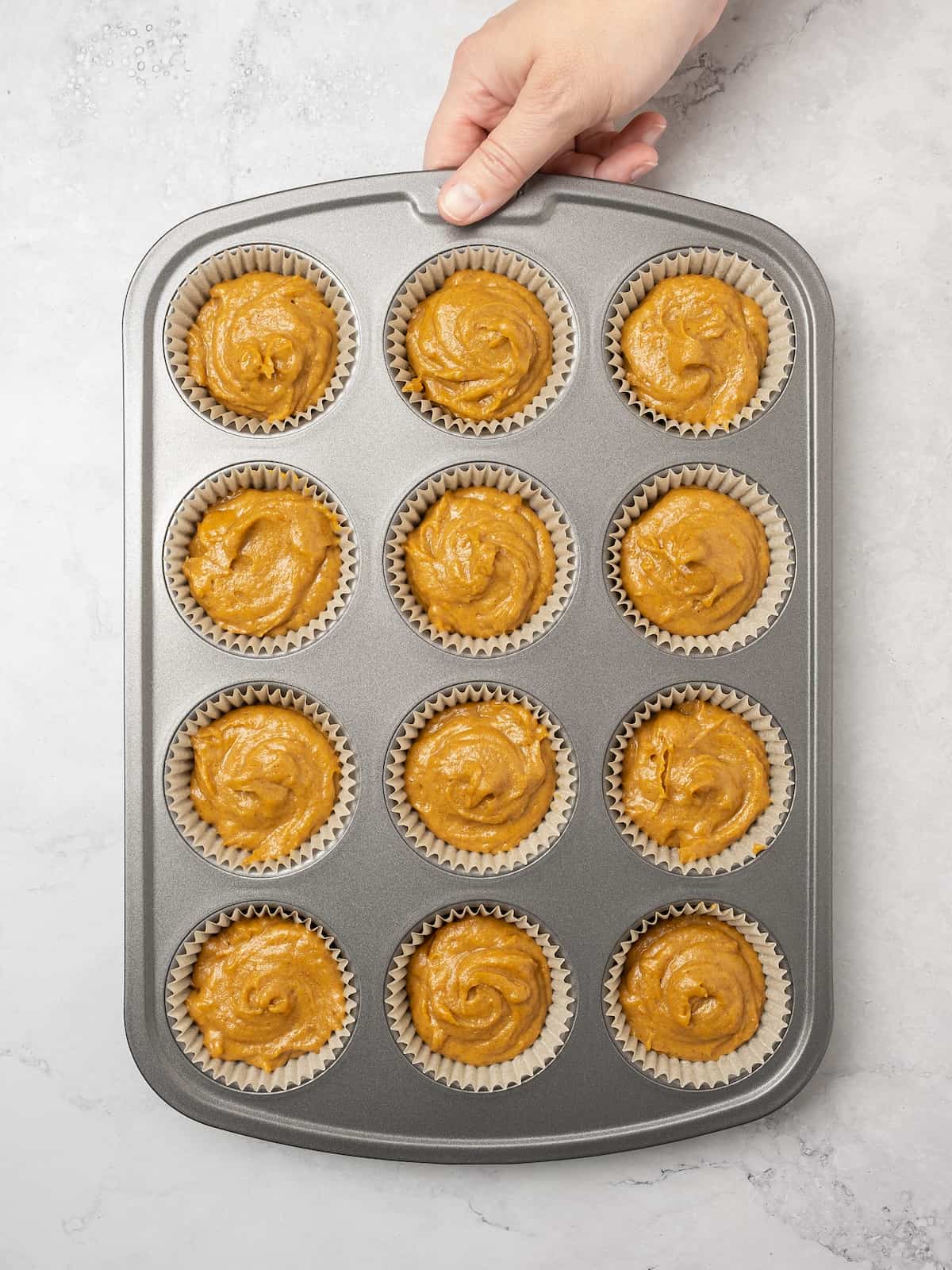 A hand holds a metal muffin tin filled with cupcake batter in cupcake liners.