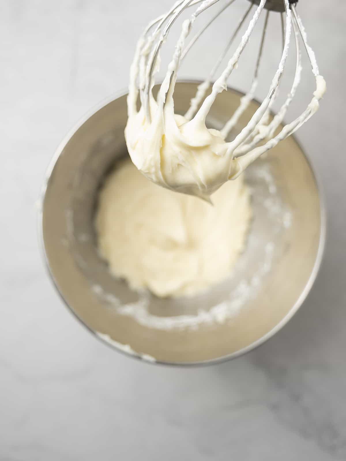 The whisk attachment of a stand mixer held above a metal bowl with beaten cream cheese frosting.