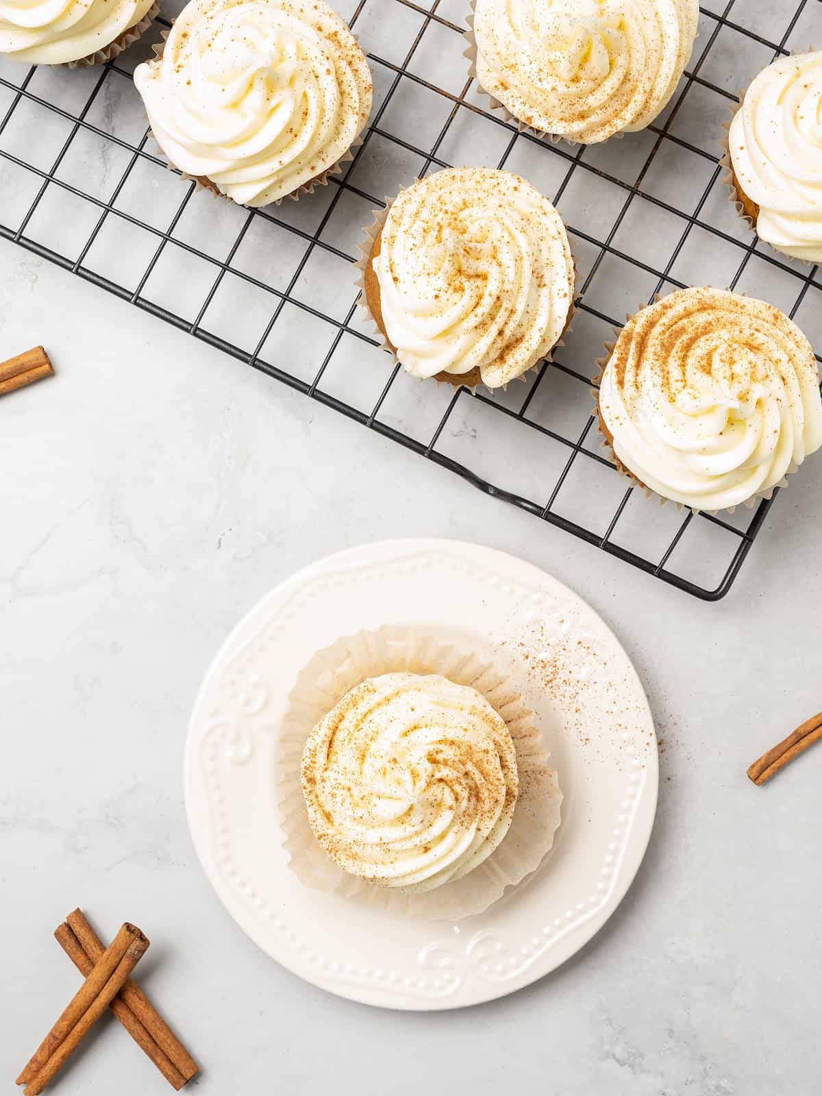 Top view of a frosted gluten free pumpkin cupcake on a plate, sprinkled with cinnamon next to more frosted cupcakes on a wire rack.