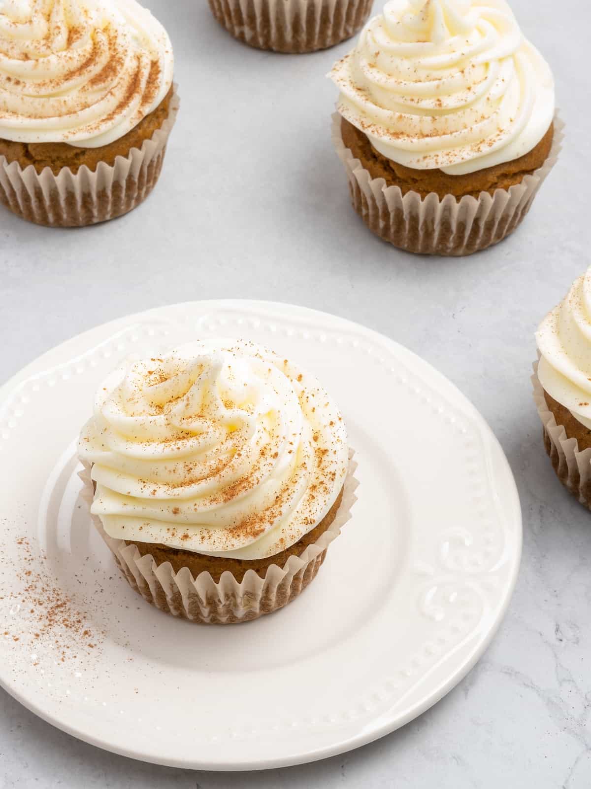 A frosted gluten free pumpkin cupcake sprinkled with cinnamon on a plate, surrounded by more frosted cupcakes.