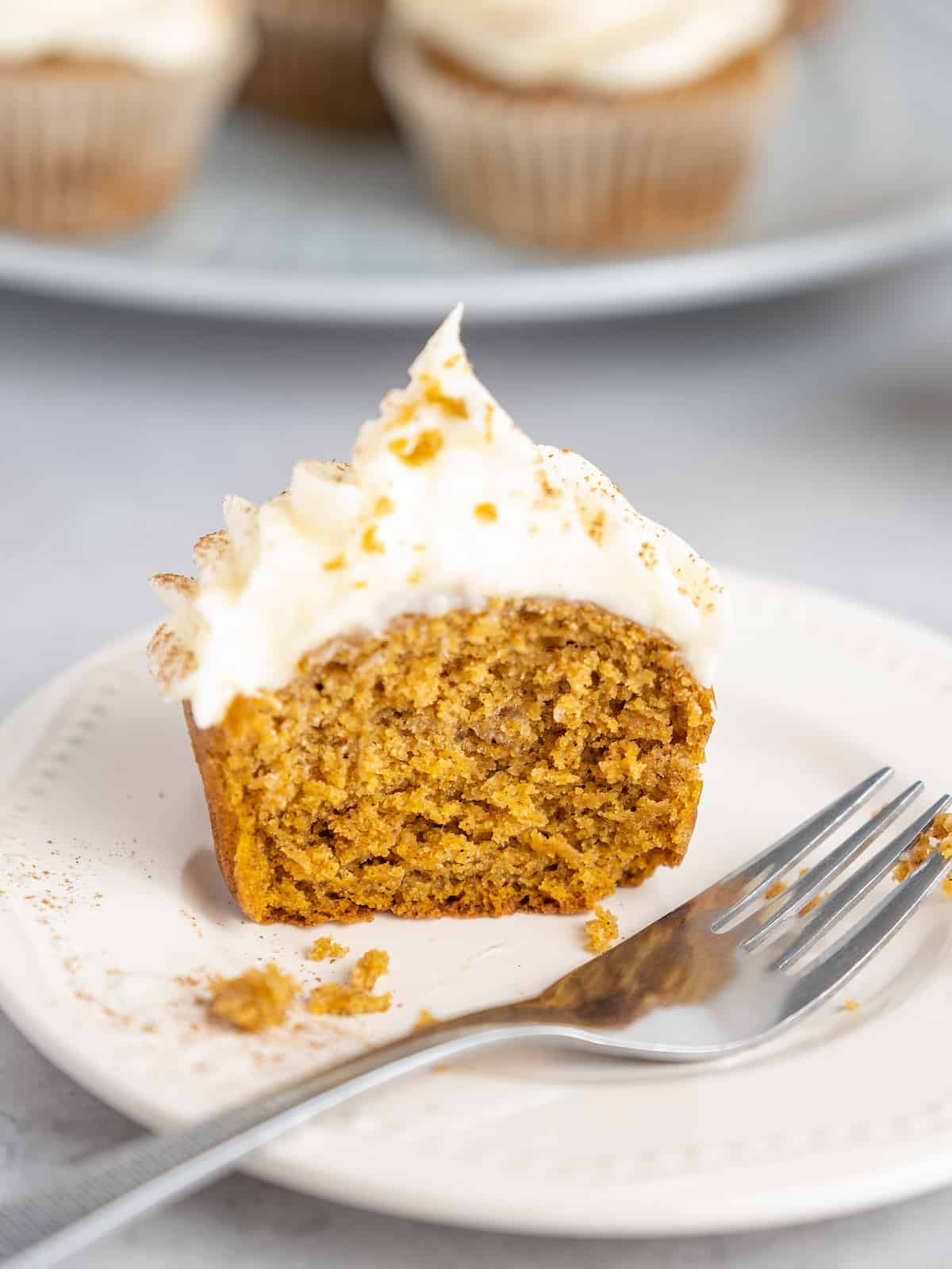 One half of a frosted pumpkin cupcake on a white plate next to a fork.