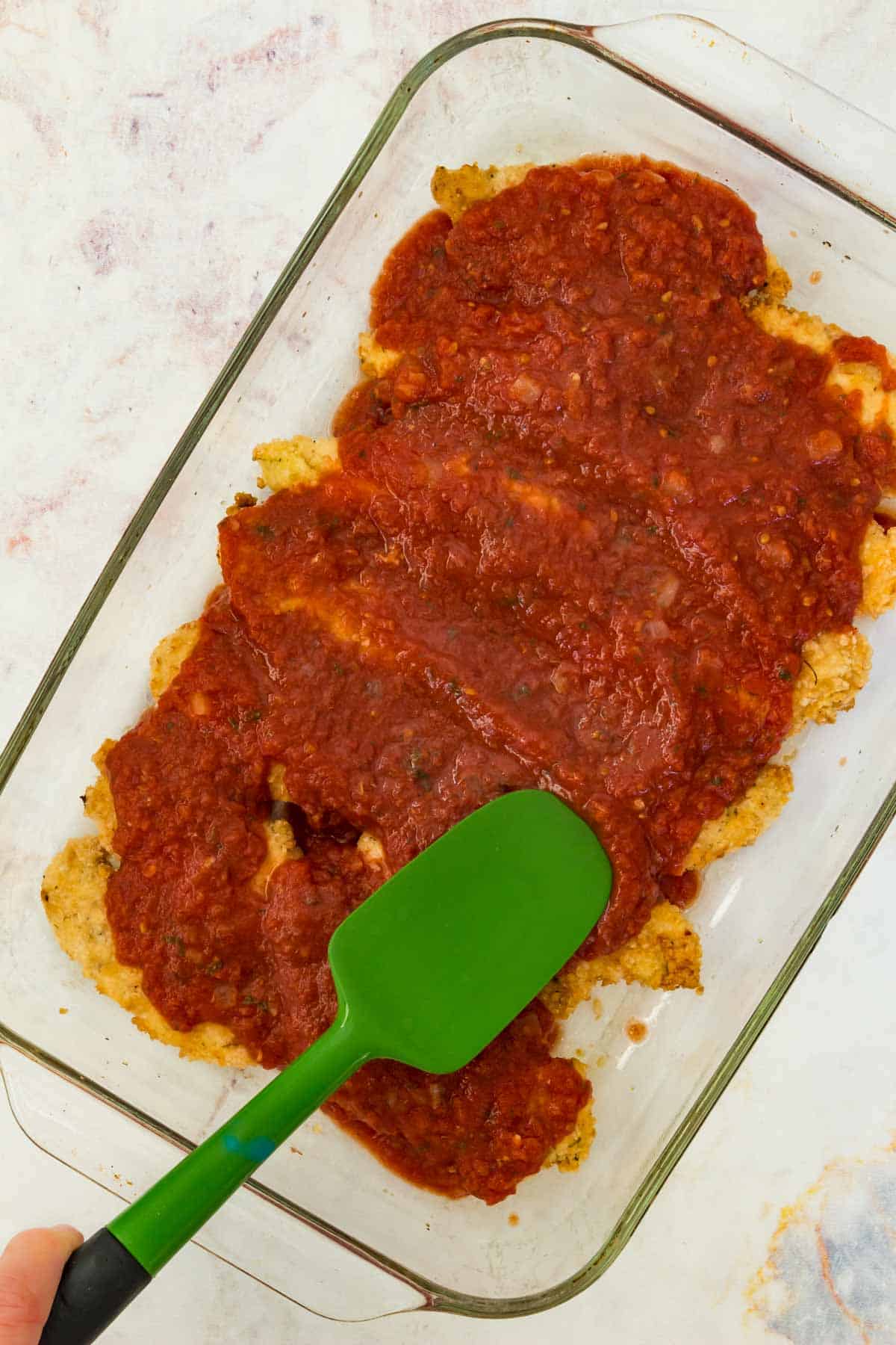 A spatula is used to spread marinara sauce overtop chicken tenders in a glass baking dish.