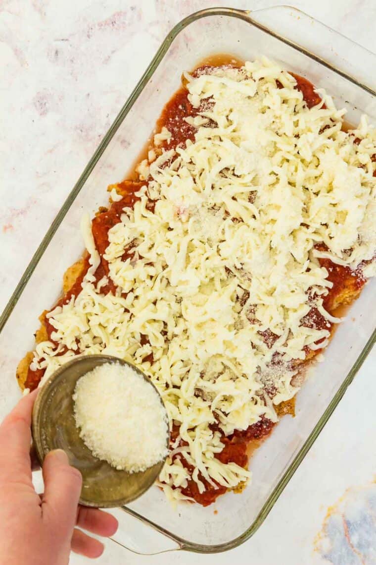 A hand holding a dish of grated parmesan cheese over chicken tenders covered in marinara in a baking dish.