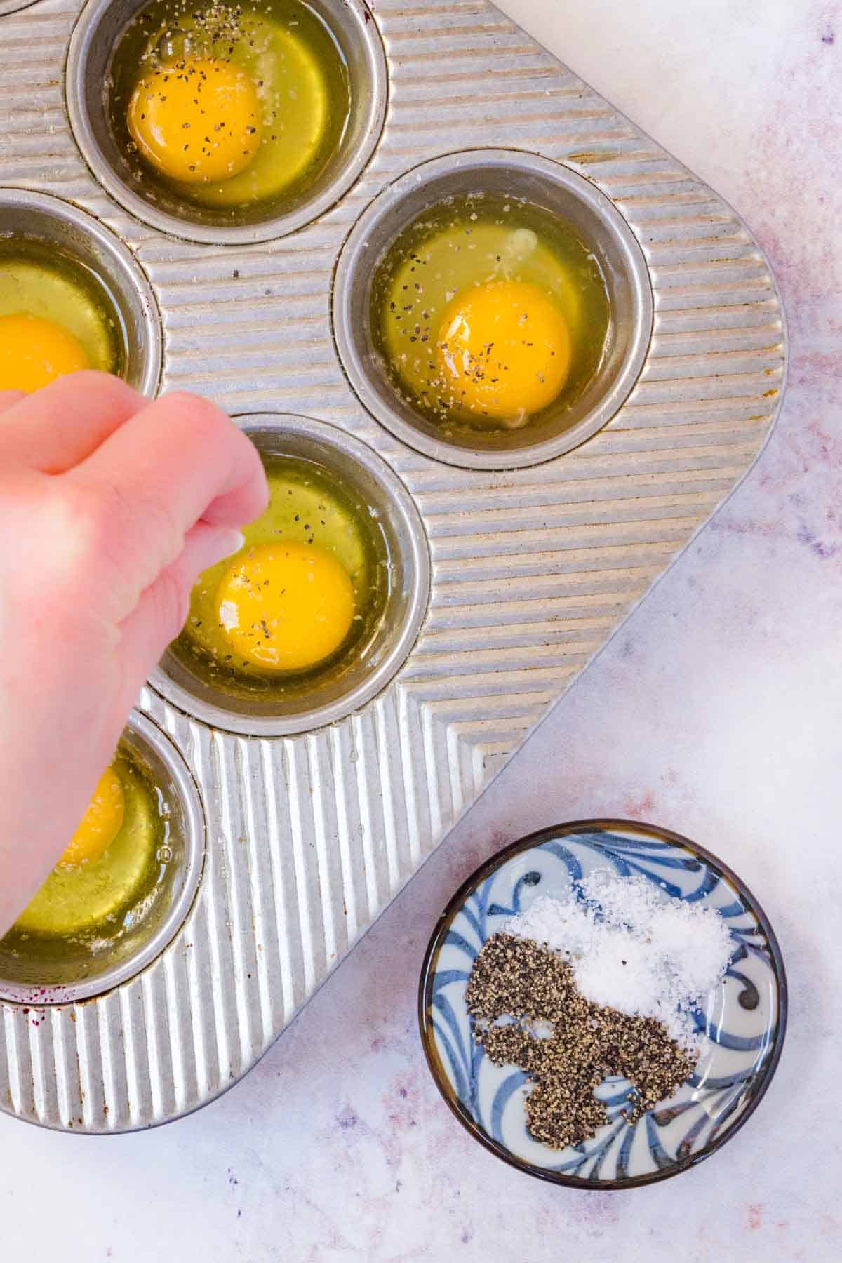 A hand sprinkles seasoning overtop of raw eggs in the wells of a metal muffin tin.