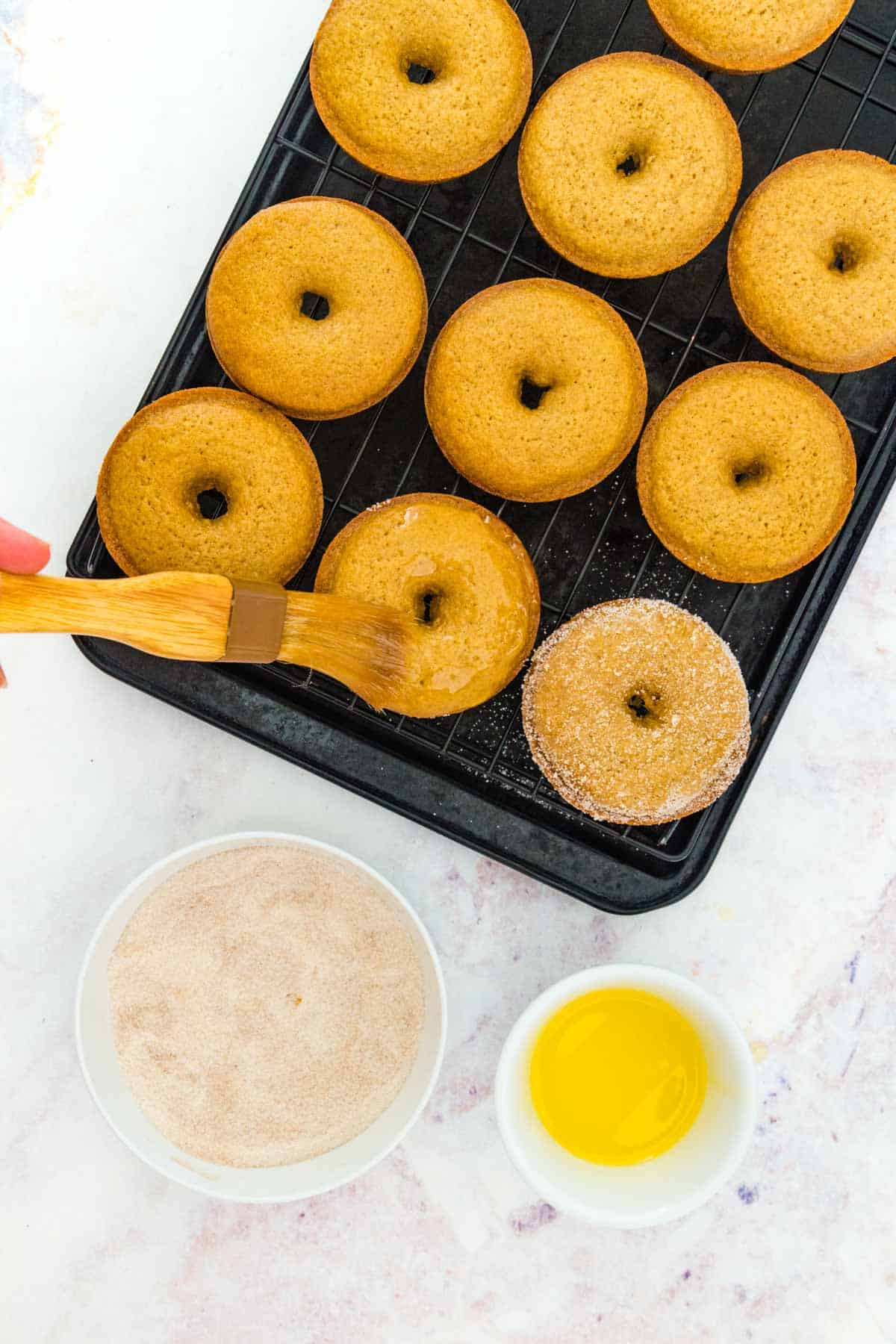 Gluten free apple cider donuts on a baking tray are brushed with melted butter, next to a small bowl of cinnamon sugar.