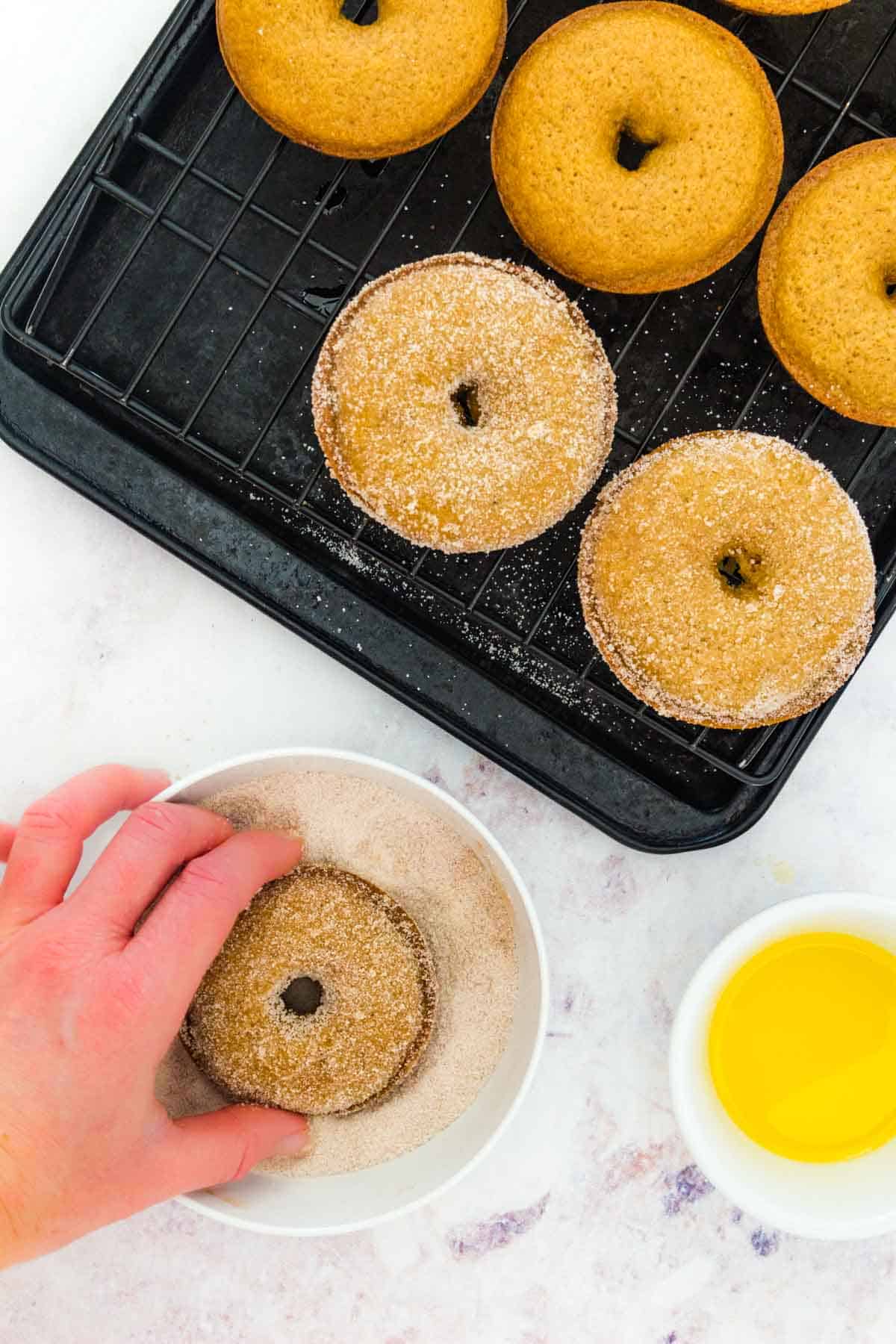 An apple cider donut is dipped into a bowl of cinnamon sugar, next to a tray of plain donuts.