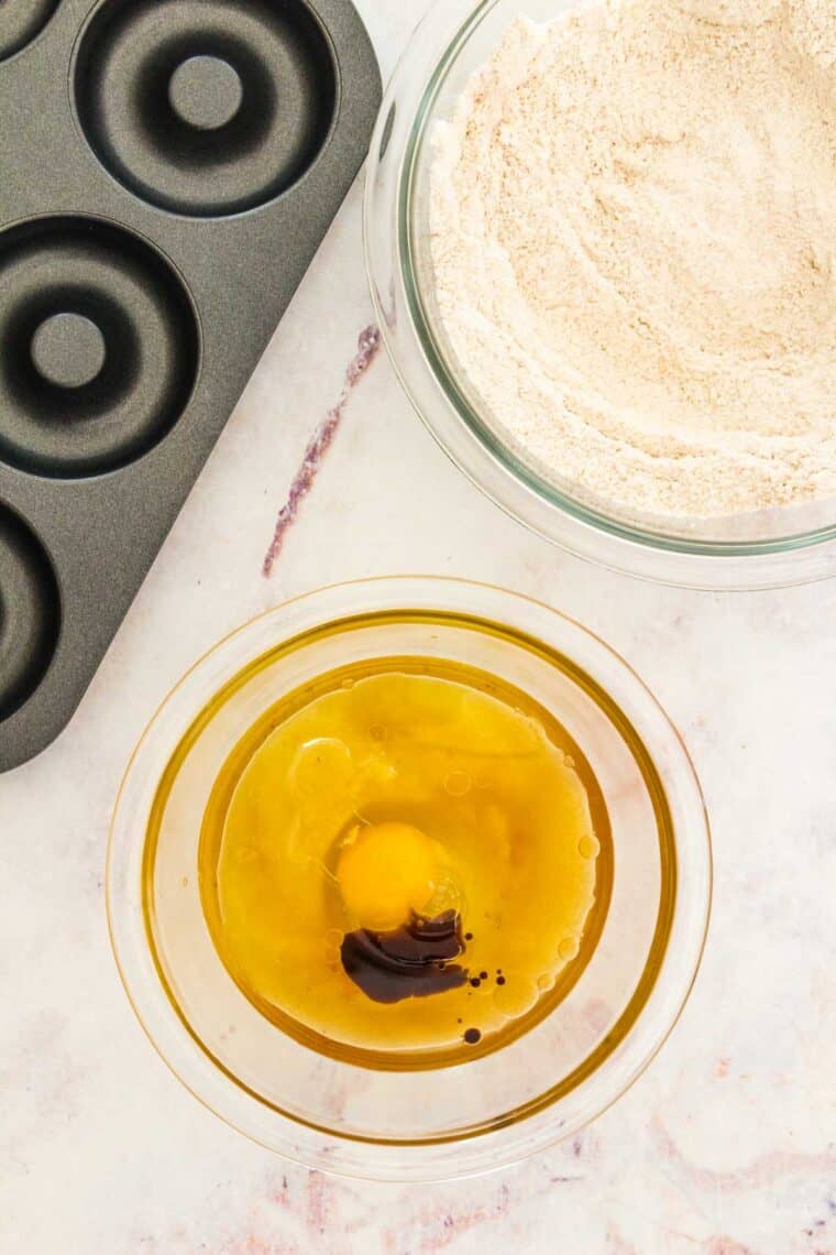 The wet ingredients for donut batter are combined in a small bowl next to a larger bowl filled with dry ingredients, next to an empty donut pan.