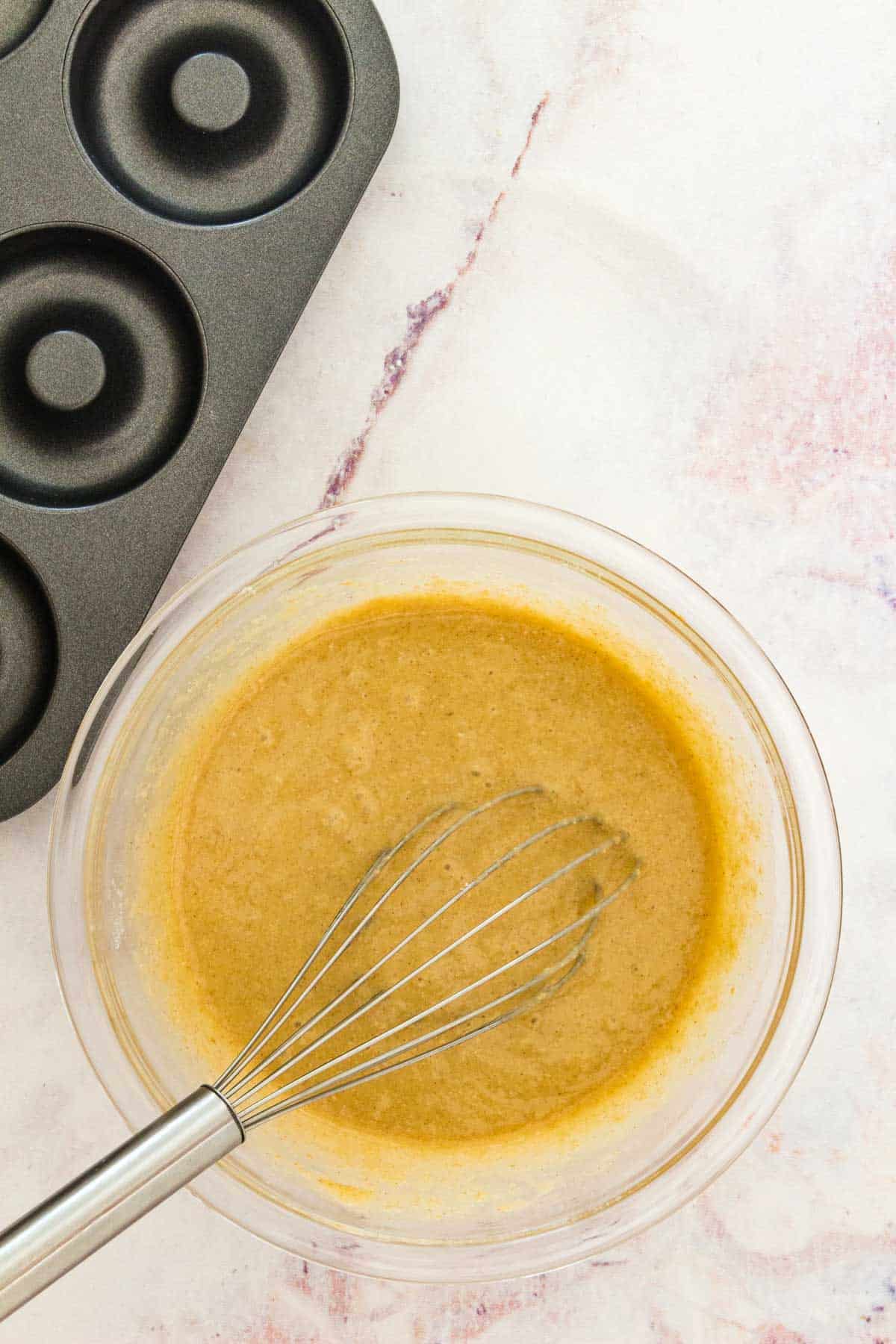 A bowl of donut batter with a whisk, next to an empty donut pan.