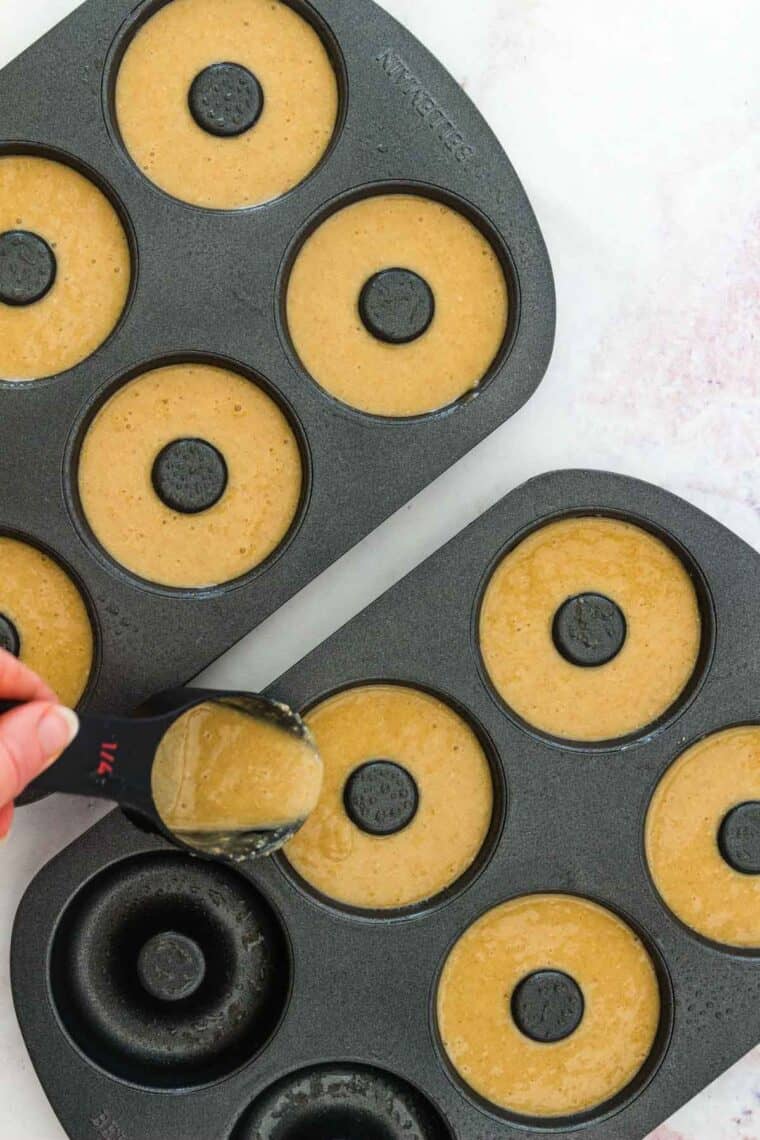 Soft, homemade Gluten Free Apple Cider Donuts are the ultimate sugary treat. These baked donuts and a cup of coffee are a cozy taste of fall! A hand uses a measuring spoon to fill the wells in a metal donut pan with batter.