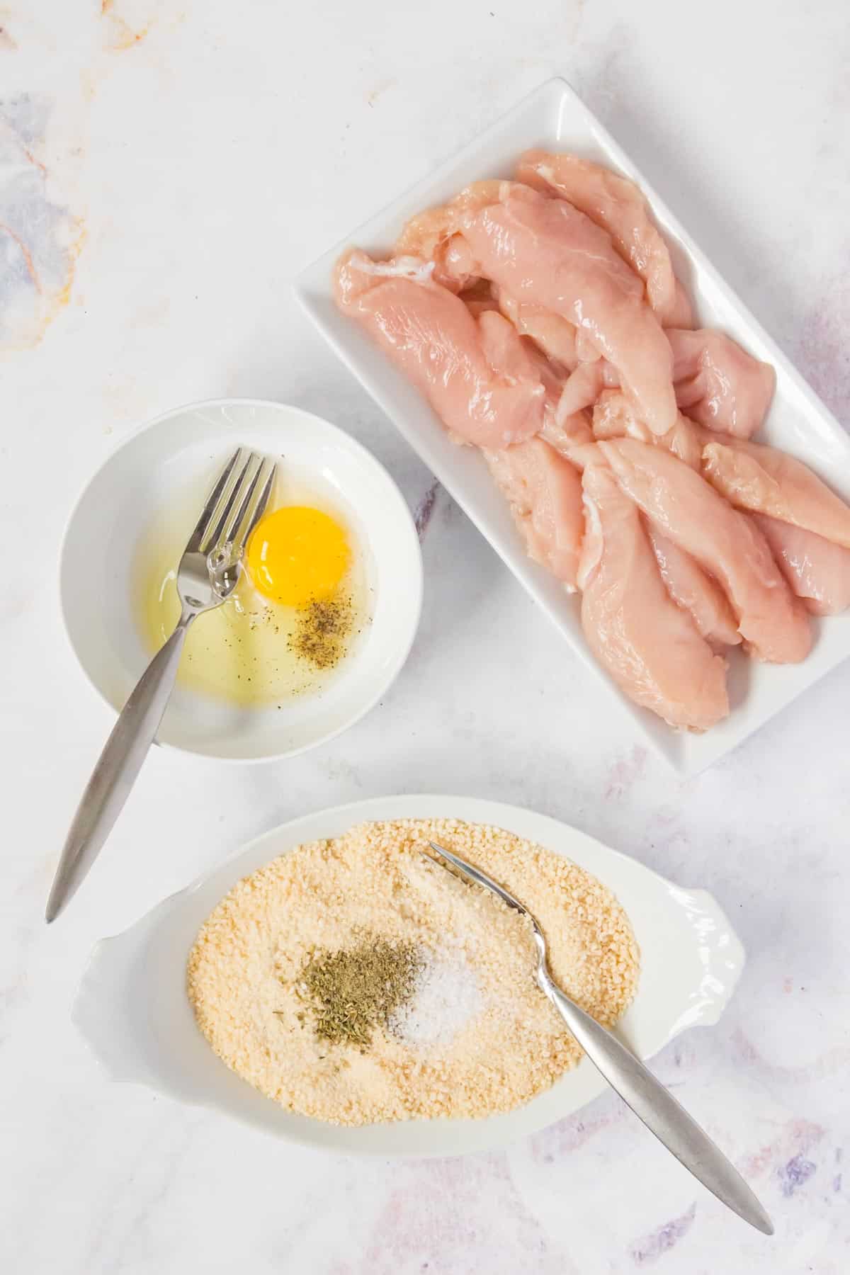 A rectangular platter of raw chicken strips, next to a dish filled with bread crumbs and seasoning, and a smaller bowl with an egg.