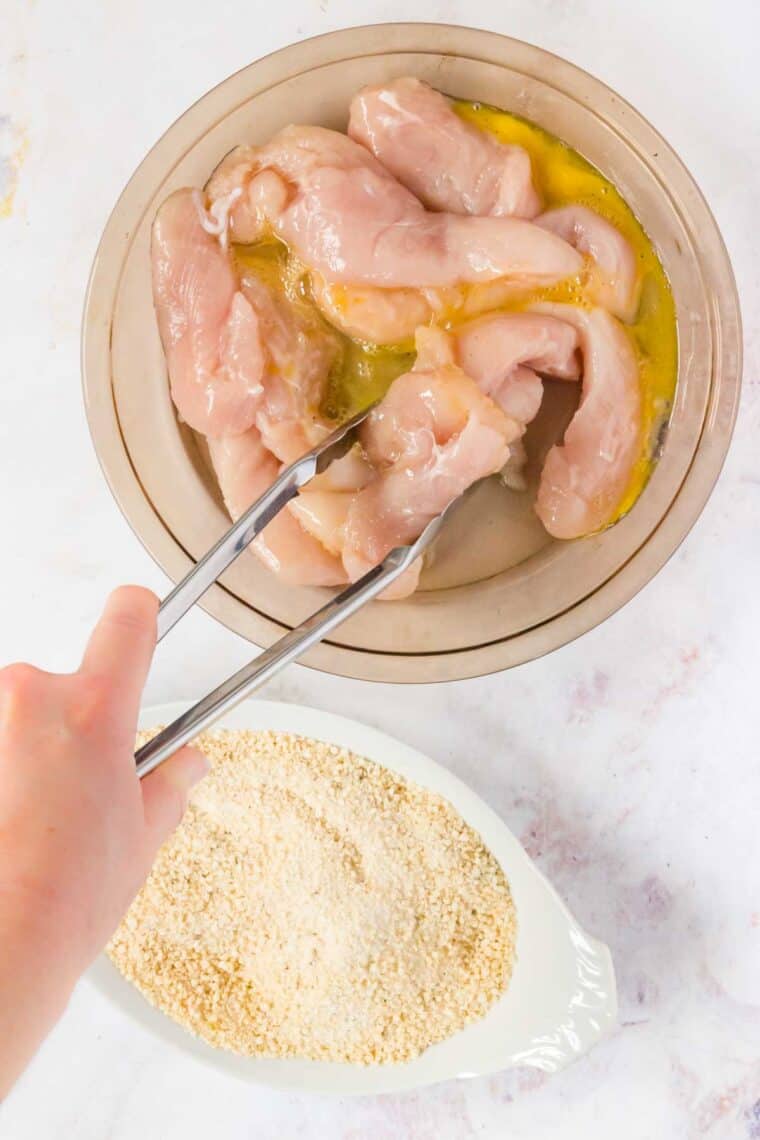 A hand uses metal tongs to toss chicken strips in a bowl of egg wash, next to a dish with seasoned bread crumbs.