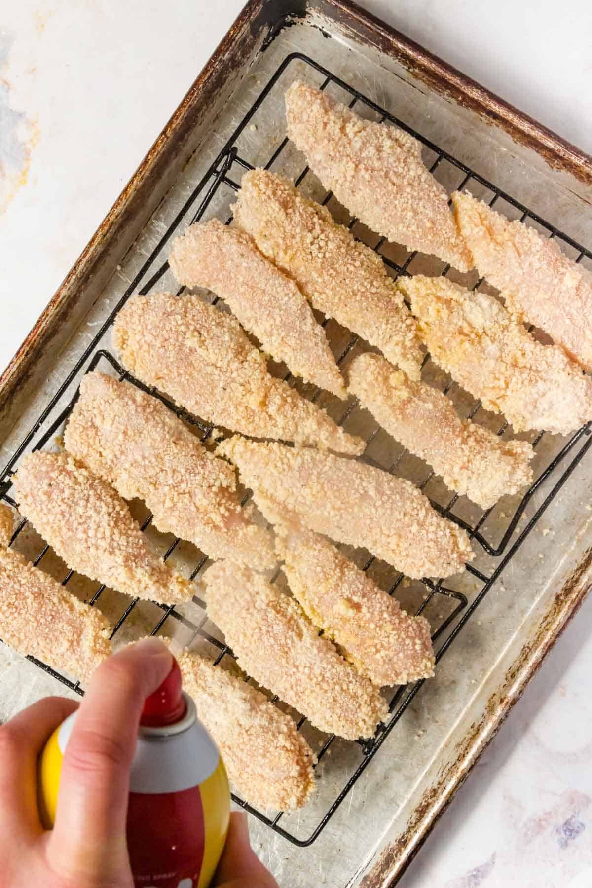A hand sprays cooking oil over unbaked chicken tenders coated in breading, lined up on a wire rack on top of a baking sheet.