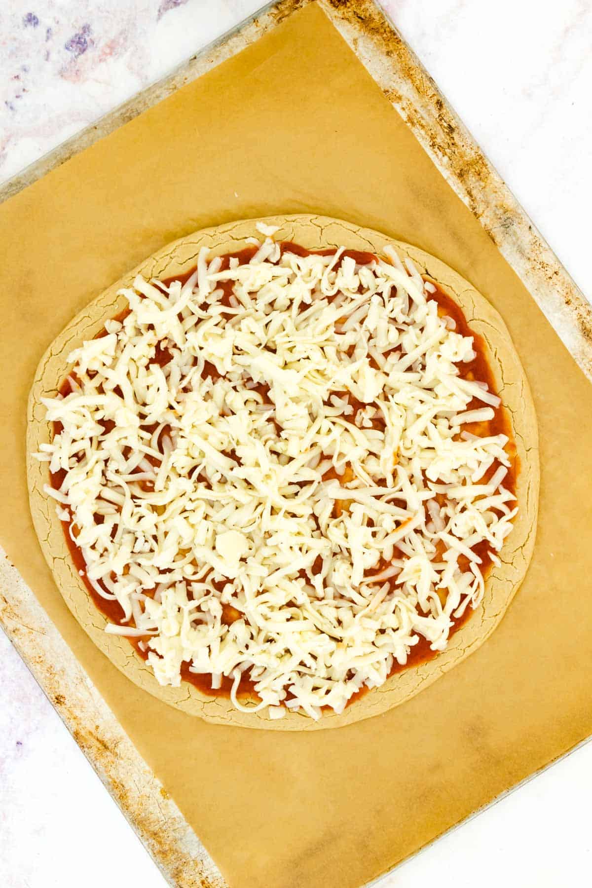 Gluten free pizza crust topped with pizza sauce and cheese on a parchment-lined baking sheet.