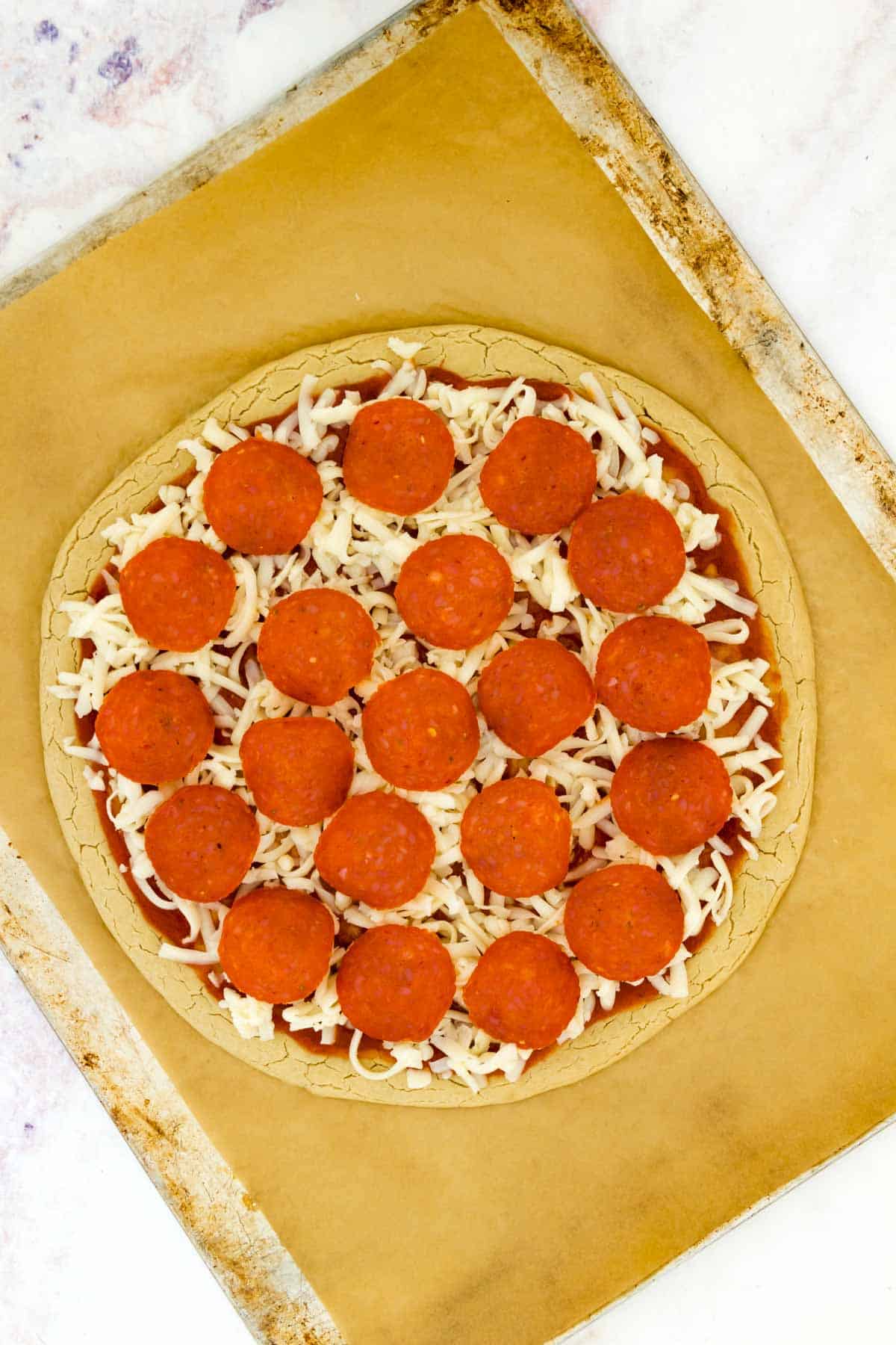 Gluten free pizza crust topped with pizza sauce, cheese, and pepperoni slices on a parchment-lined baking sheet.