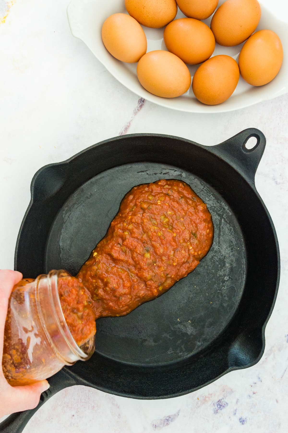 Salsa being poured into a cast iron skillet beside a bowl full of whole raw eggs.