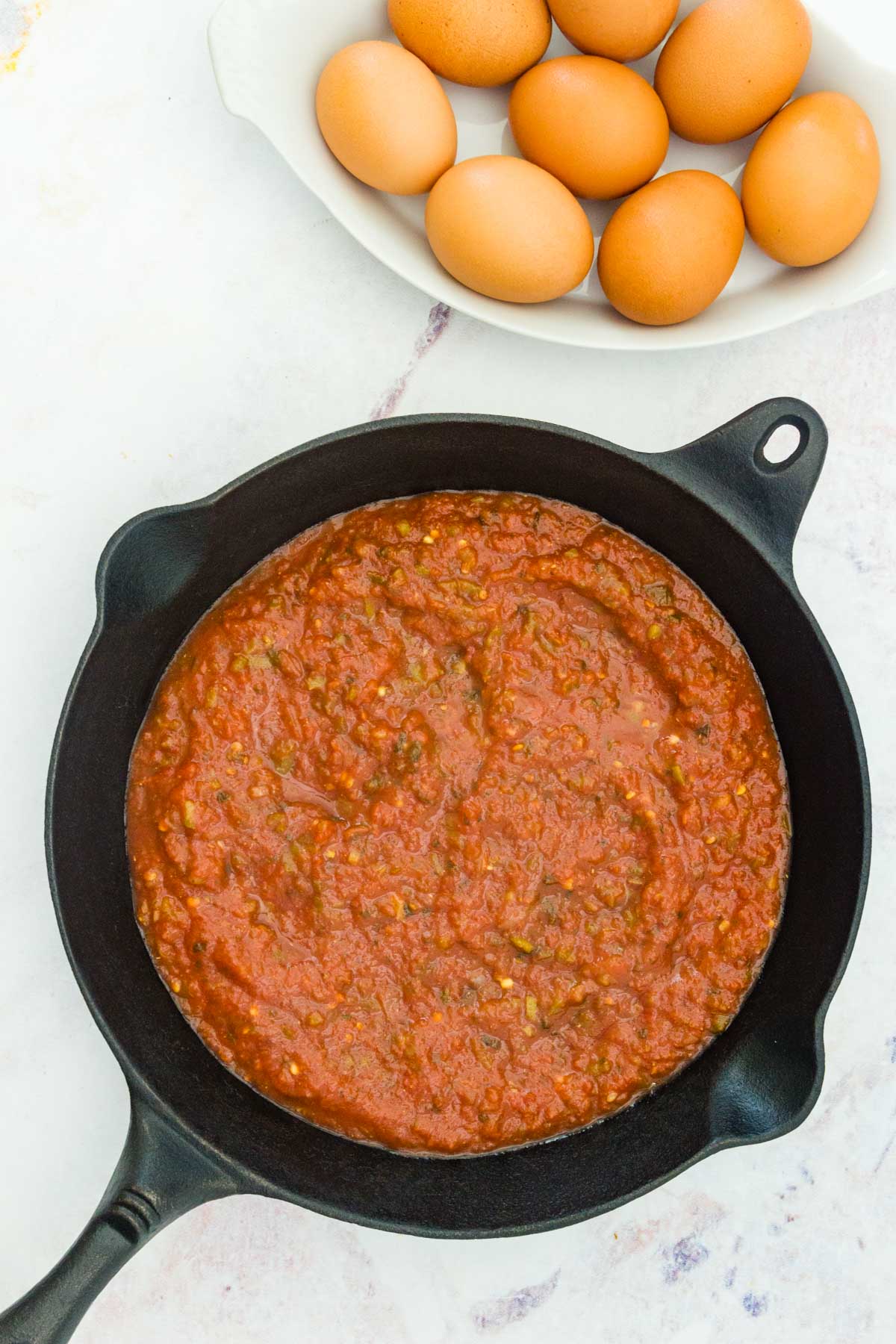 Salsa spread evenly across the bottom of a skillet next to a bowl of 8 eggs.