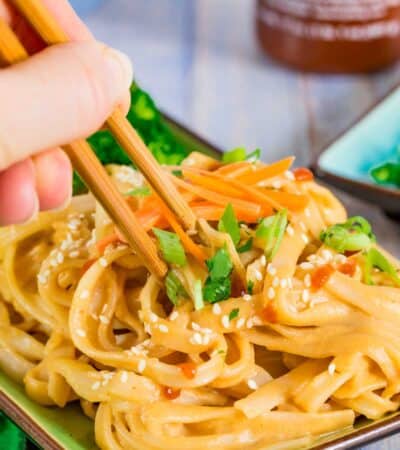 Spicy Sesame Noodles with vegetables for garnish being picked up by a pair of chopsticks.