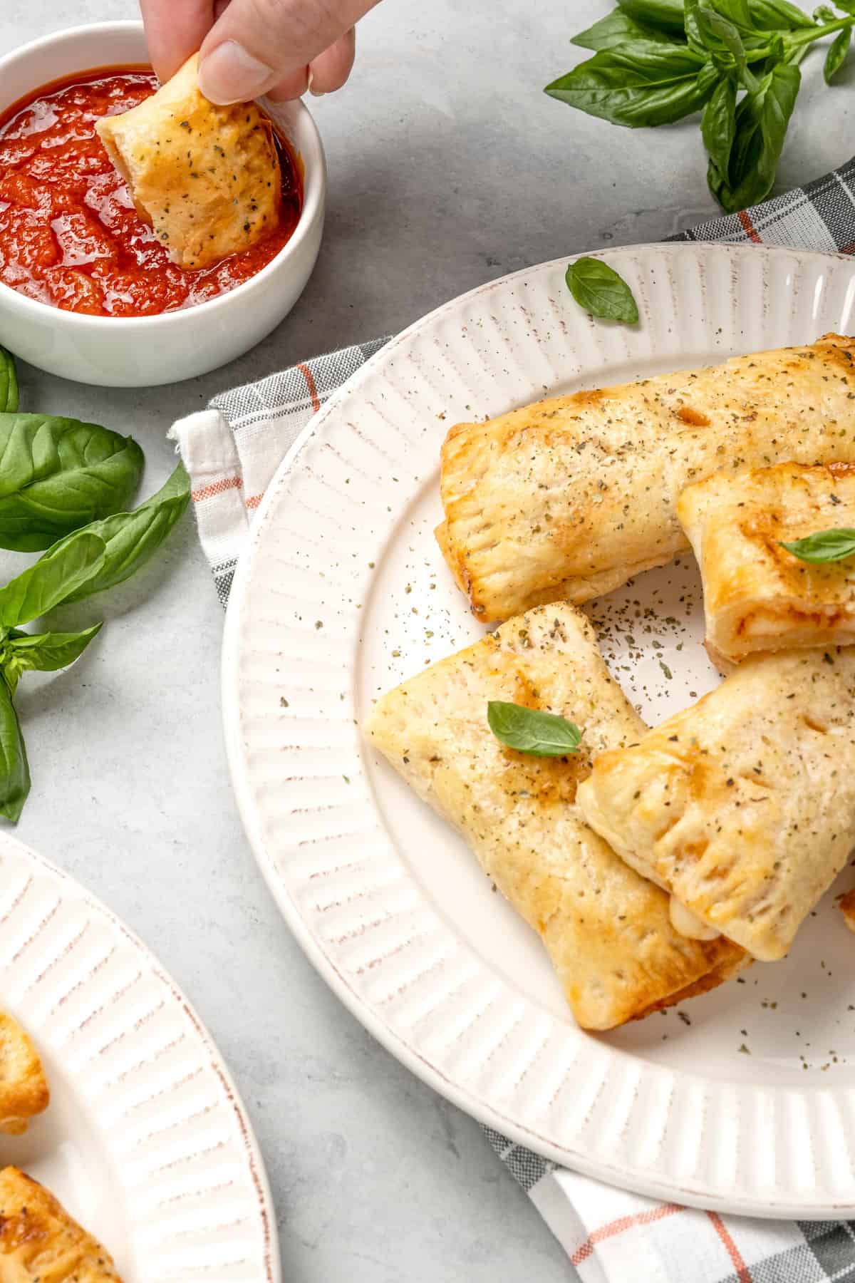 Air fryer gluten free pizza rolls on a white plate, garnished with basil leaves, with a pizza roll being dipping in marinara sauce in the background.