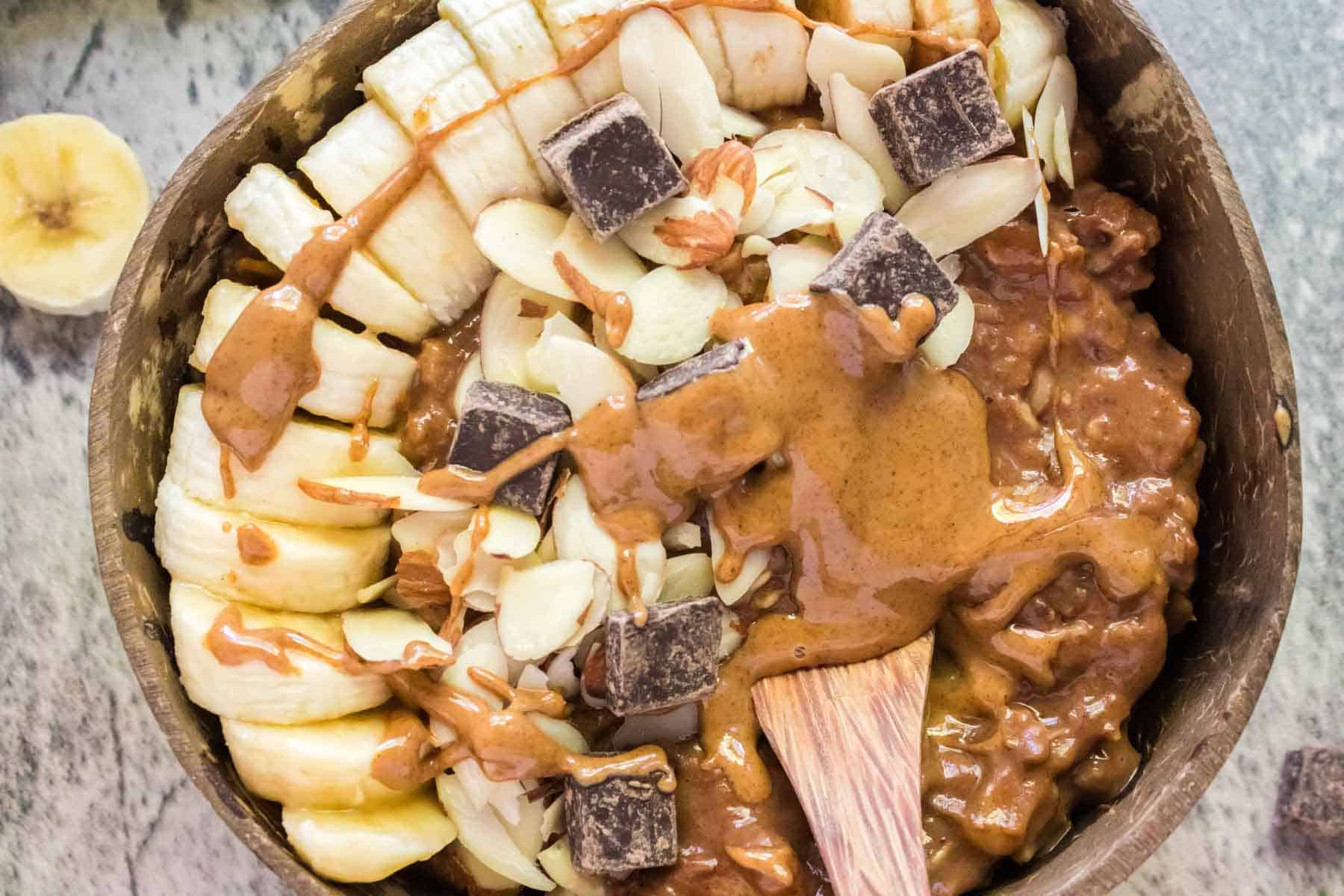 A close-up of a bowl of chocolate oatmeal with banana, chocolate, nuts and nut butter for toppings.