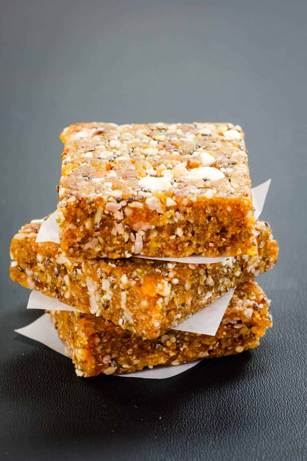 Three apricot chia energy bars stacked with squares of wax paper between each one on a black surface.