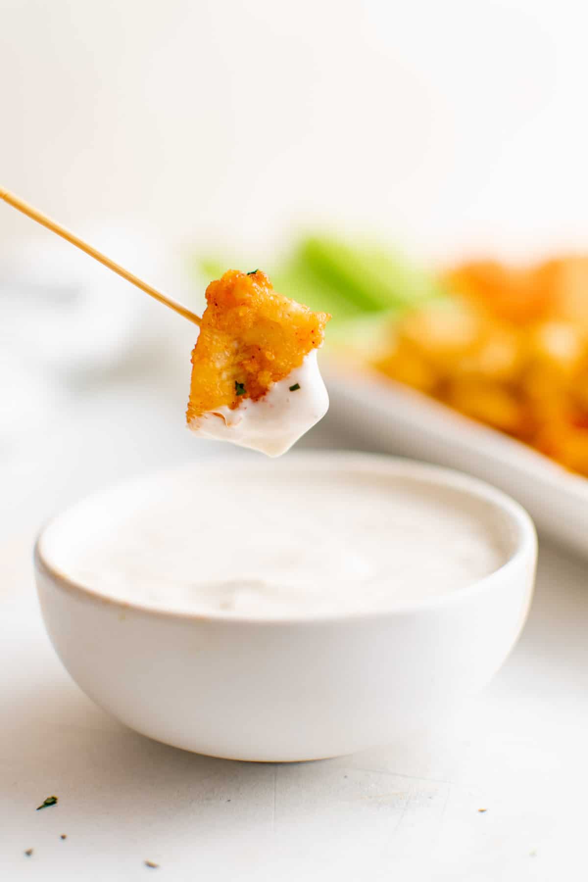 A buffalo chicken bite on the end of a toothpick is dipped into a white dipping sauce.