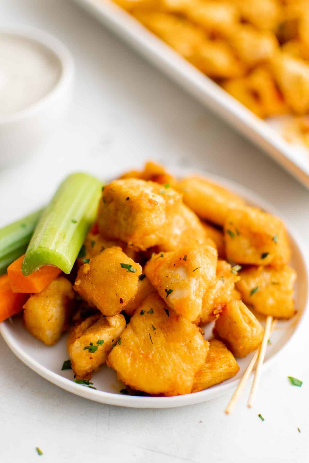 Buffalo chicken bites in a pile on a white plate, next to carrot and celery sticks.