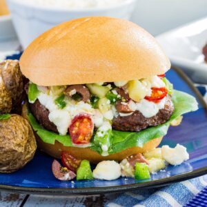 A grilled burger on a bun with tzatziki sauce and a Greek salad topping overflowing out of the bun.