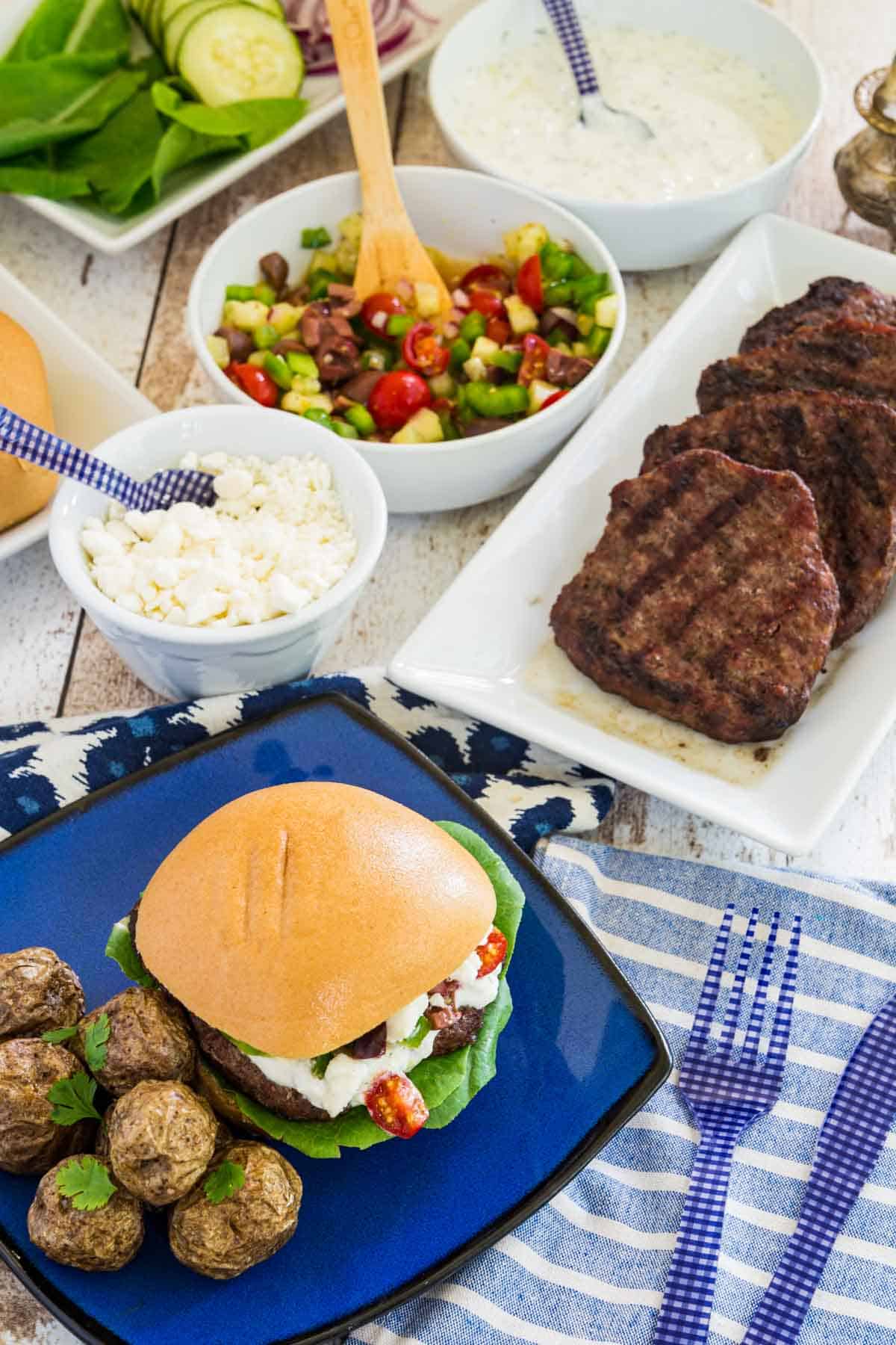 A table set with bowls and platters of burger toppings with one assembled burger on a blue plate with potatoes.