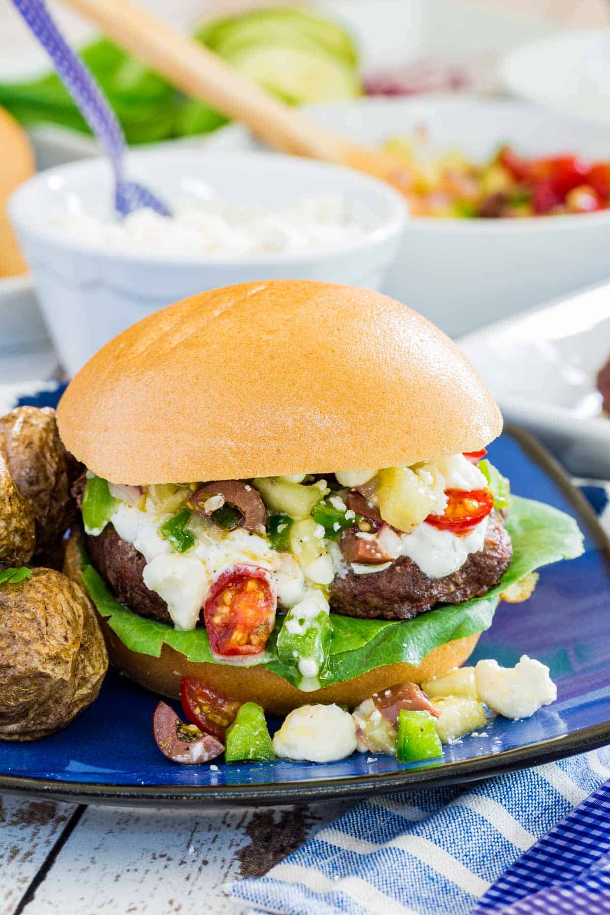 A Greek burger with tzatziki sauce and Greek salad topping dripping off of it onto a blue plate.