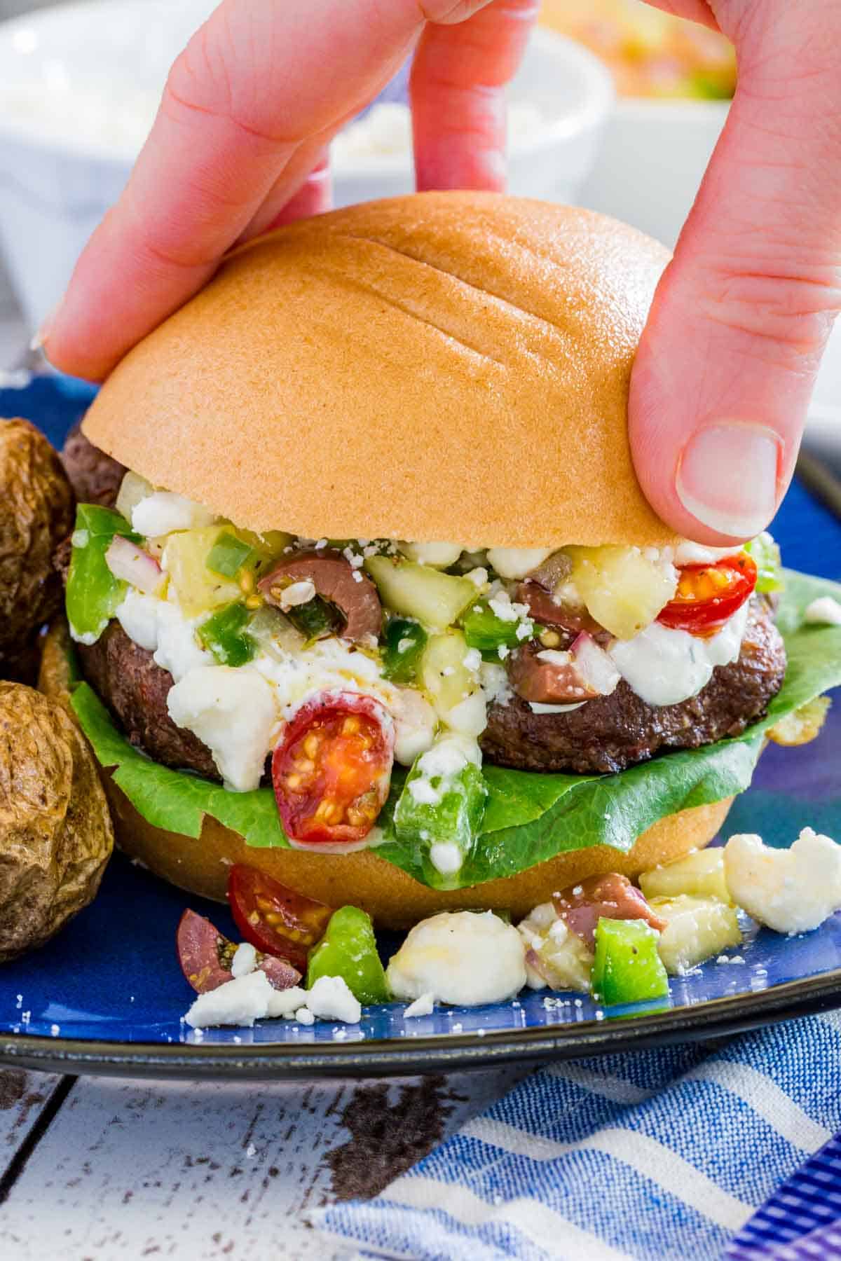 A hand putting a the top of a bun on a burger with tzatziki and Greek salad topping.