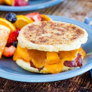 English muffin breakfast sandwich with bacon, egg, and cheese on a blue plate with fruit.