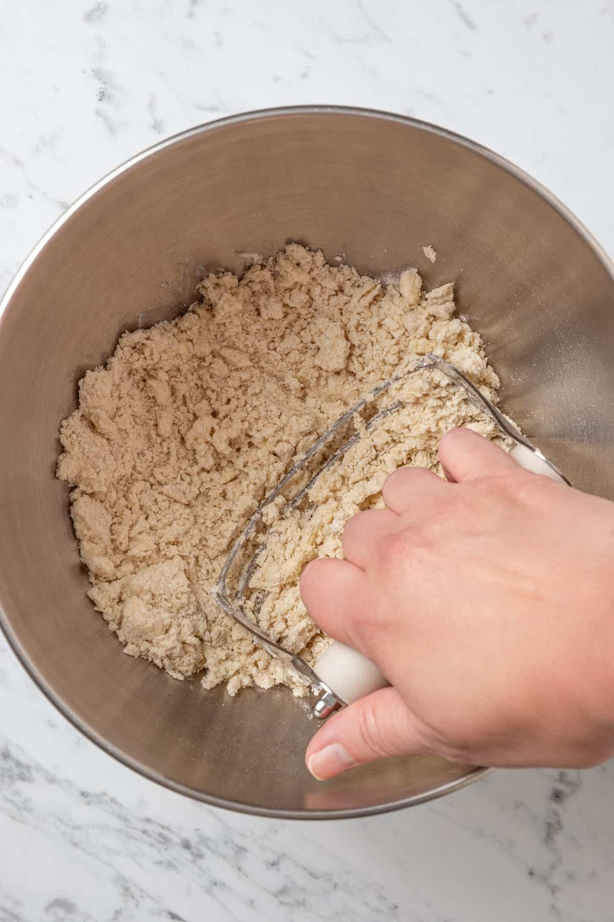 A hand uses a pastry cutter to cut butter into the dry dough ingredients in a mixing bowl.