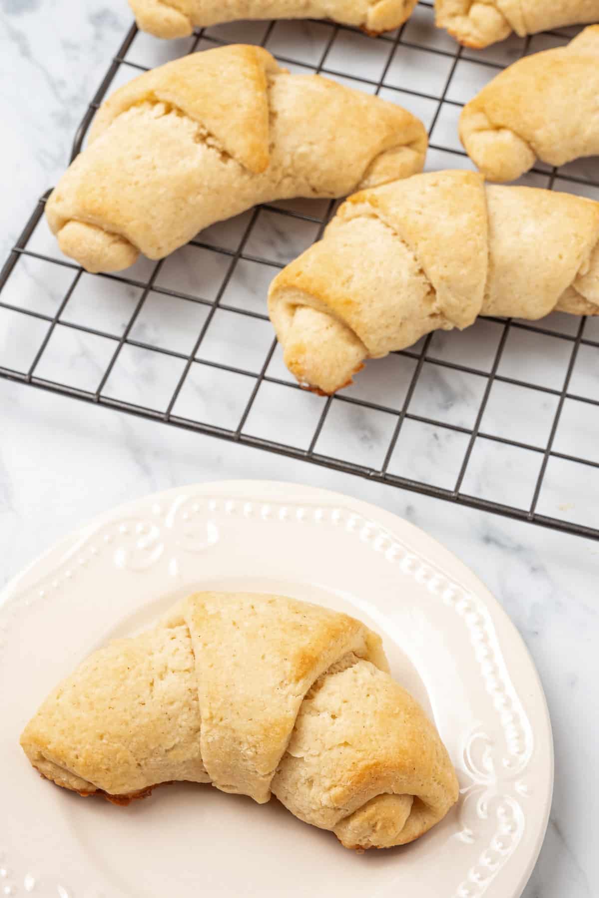 A gluten free crescent roll on a white plate next to more rolls on a wire cooling rack.