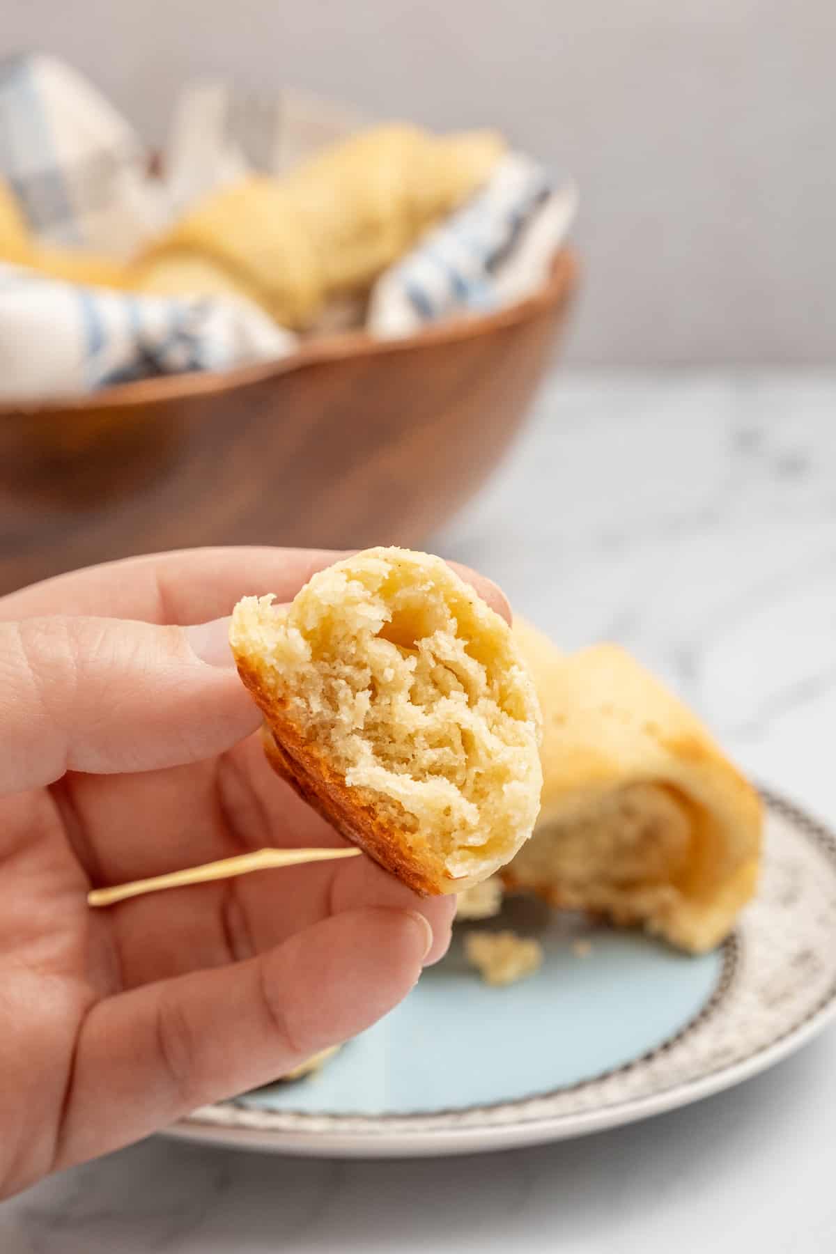 A hand holds up a piece of a soft, fluffy gluten free crescent roll in front of a bowl of crescent rolls in the background.