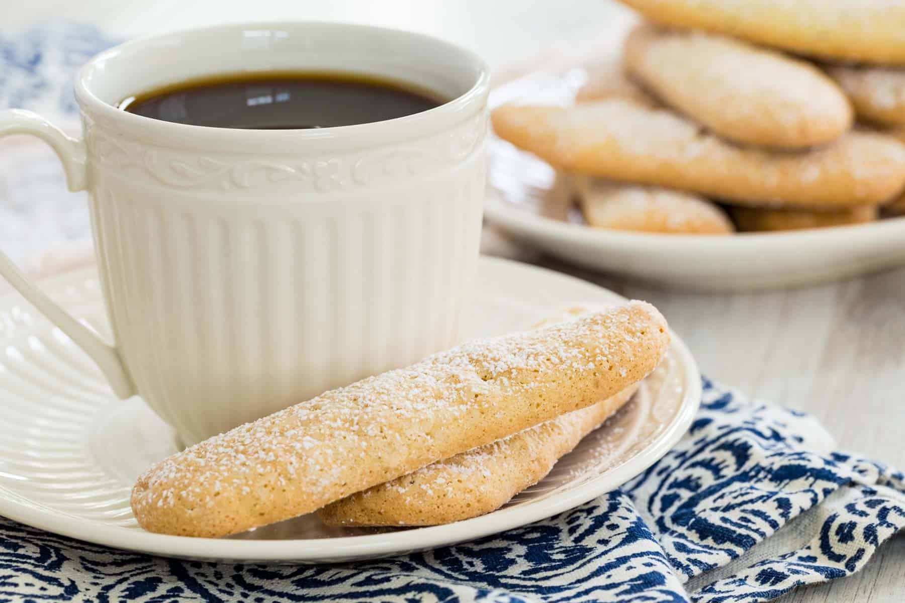 Two gluten free ladyfingers on a saucer next to a cup of coffee, with a plate of ladyfingers in the background.